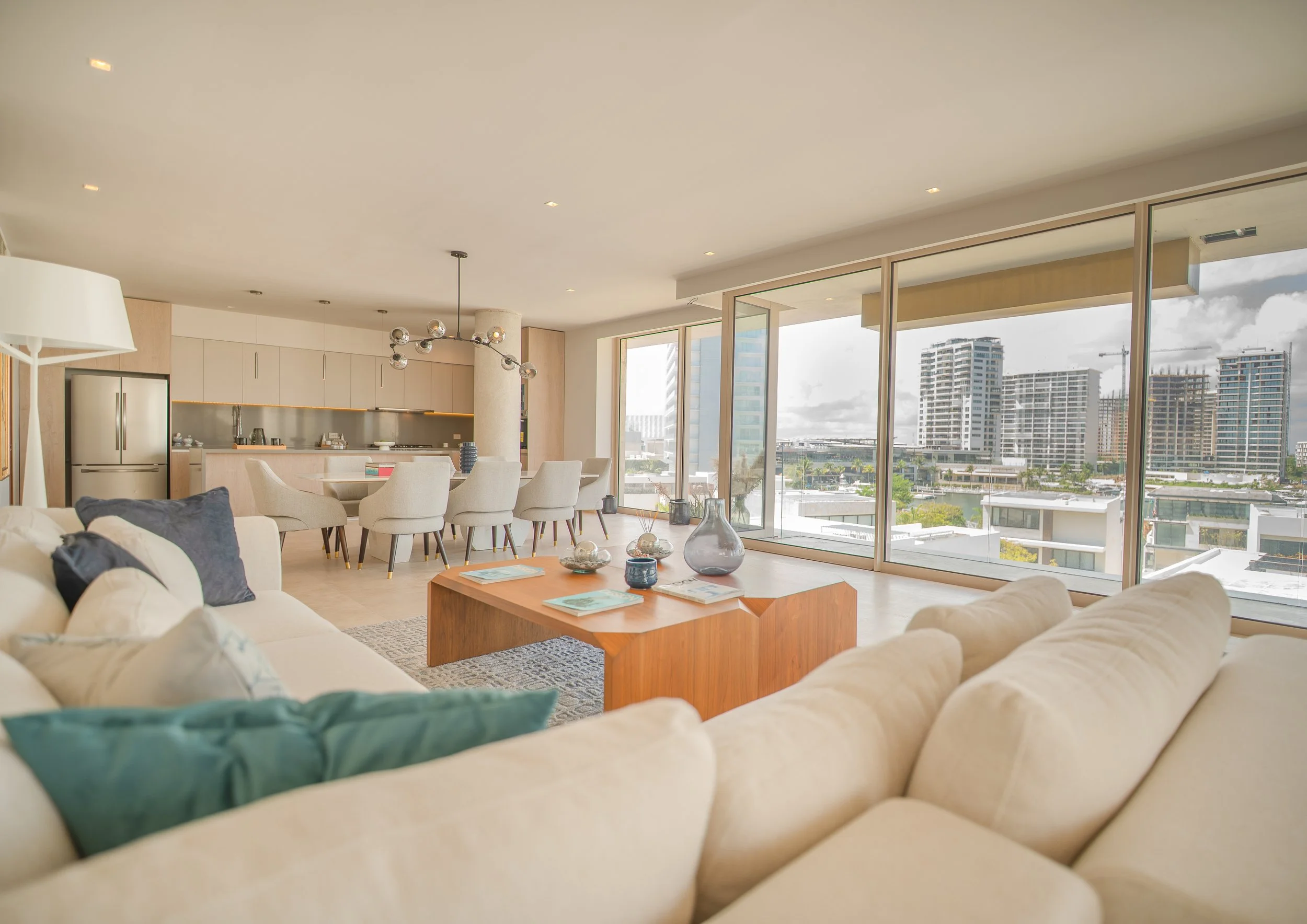 Living room with cream sofa, pillows, wooden coffee table with decorative vases, and large windows showing city buildings and a partly cloudy sky.