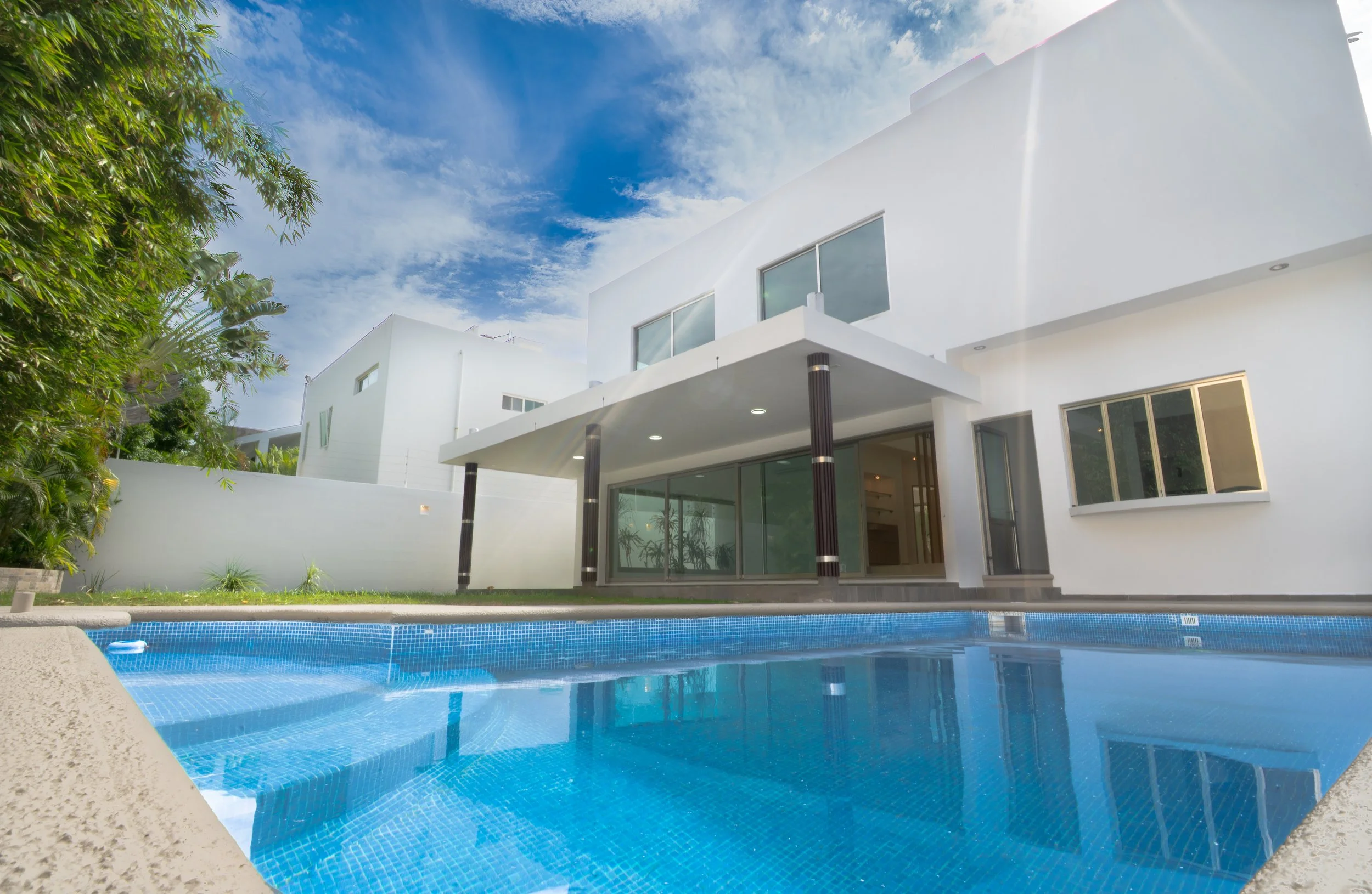 Modern white house with large windows and a balcony, surrounded by trees, with a swimming pool in the foreground under a partly cloudy sky.