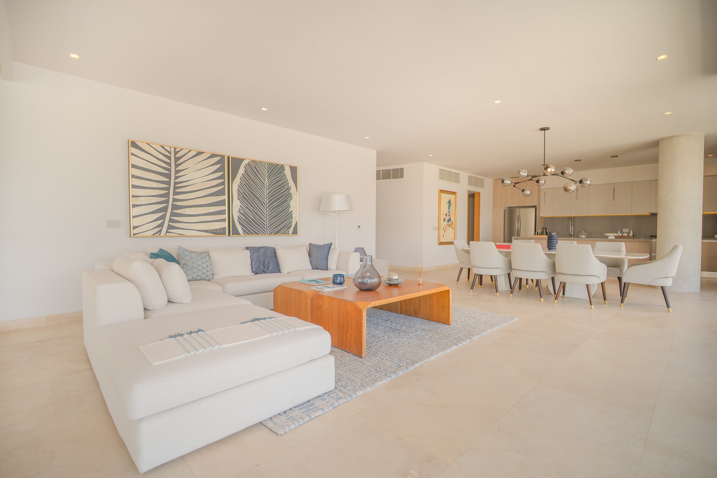 Modern open-plan living room and kitchen with a white sectional sofa, wood coffee table, dining area, contemporary lighting, and botanical wall art