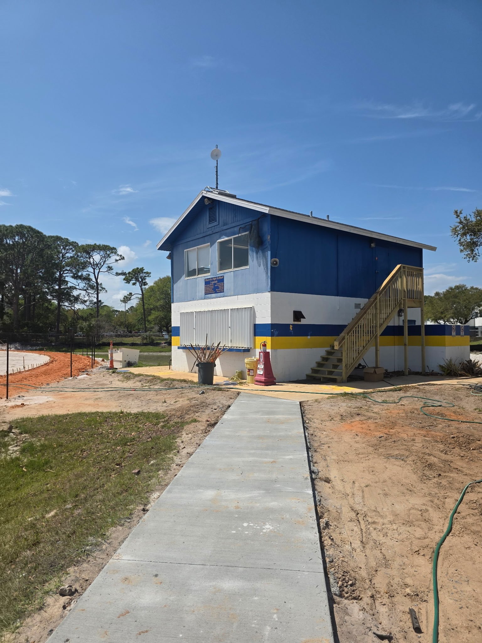 Newly constructed building with blue and white exterior, stairs on the right, and a sidewalk in the foreground. Construction materials and equipment are visible around the building, with trees and a clear sky in the background.