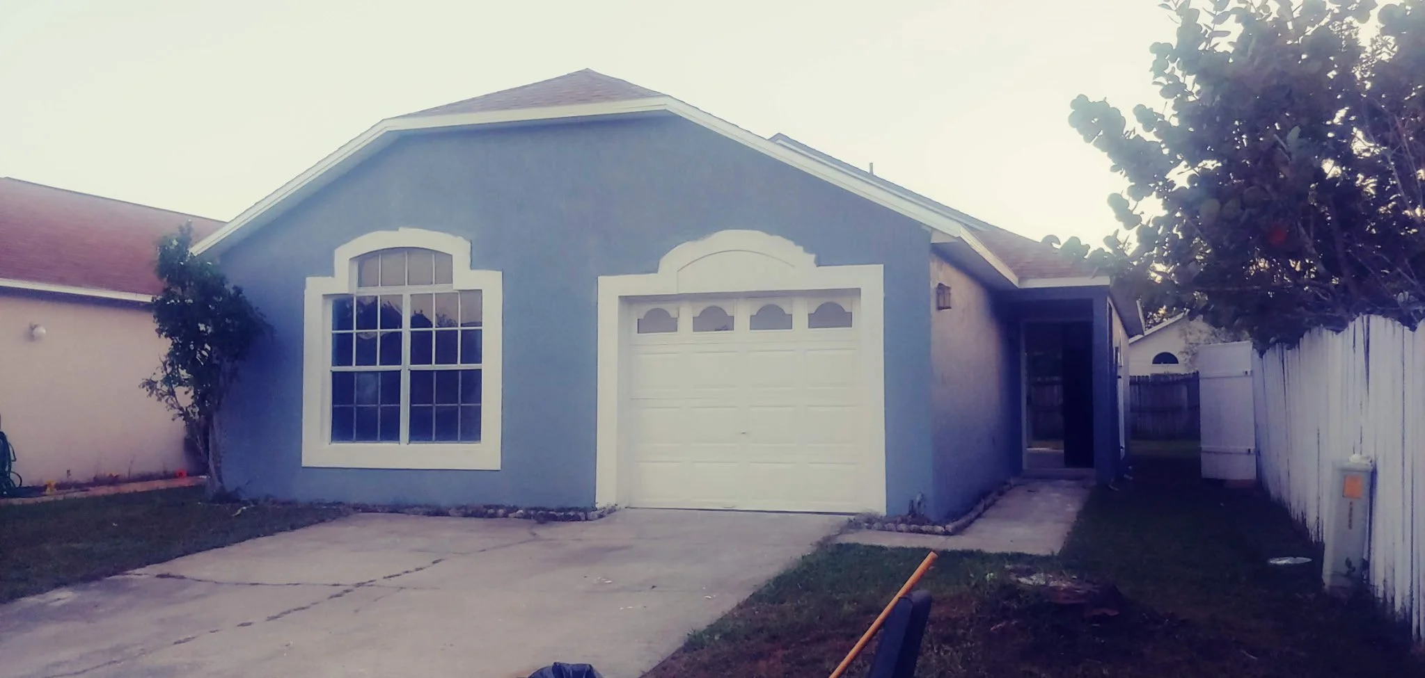 A blue house with a white garage door, large window, and a small front yard. There is a driveway and a tree on the right side. The house has a peaked roof.