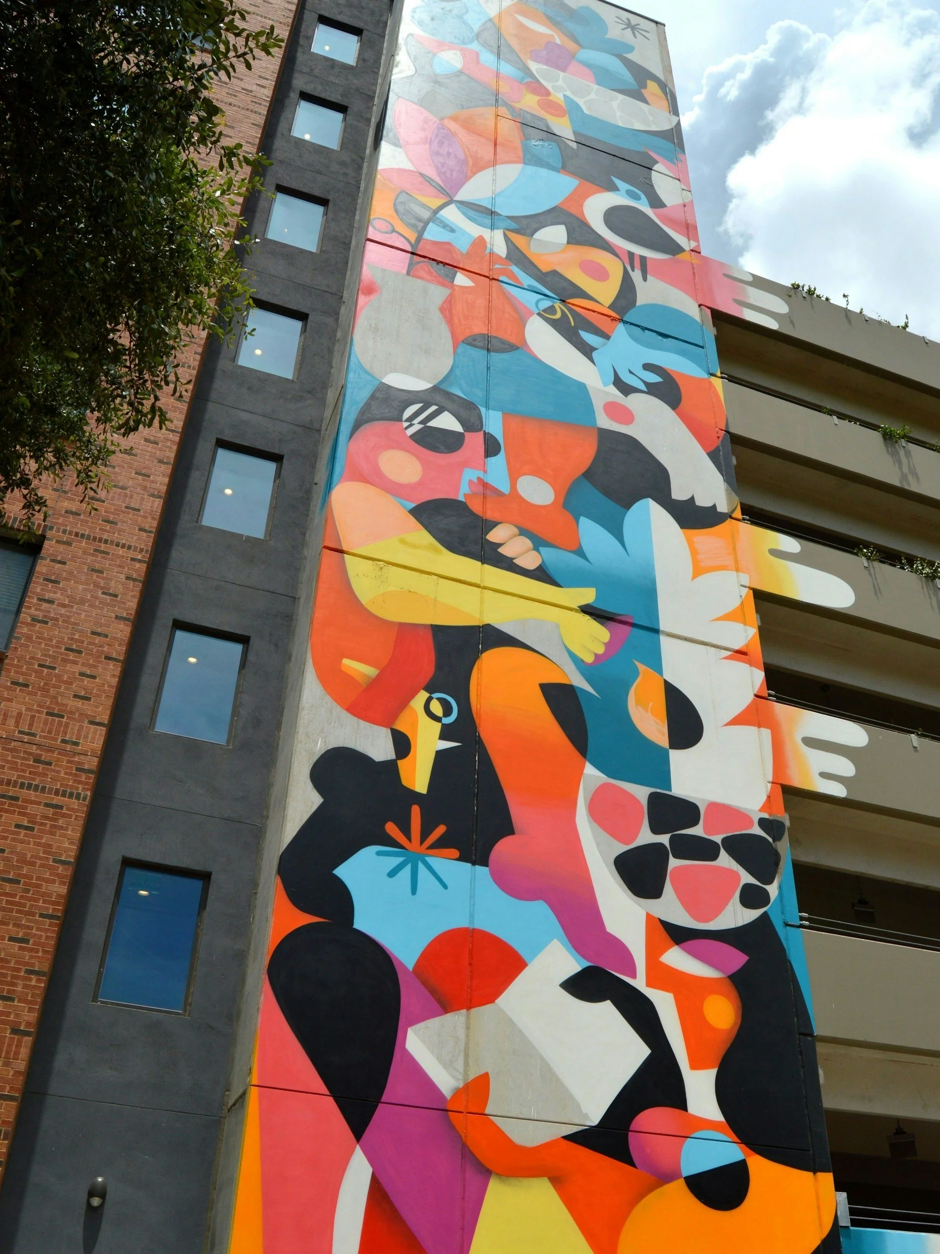 A tall building with a colorful abstract mural on its exterior, featuring various shapes and patterns in bright colors like pink, blue, orange, yellow, and black. The building has several rectangular windows and is next to a tree with green leaves. The sky is partly cloudy with blue sky showing.