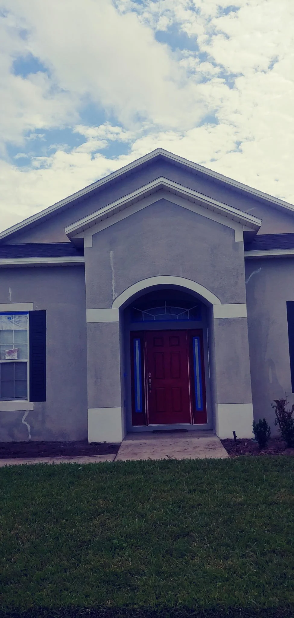 Front view of a house with a purple exterior, arched doorway with a red door, blue side panels, a window with shutters, and a lawn