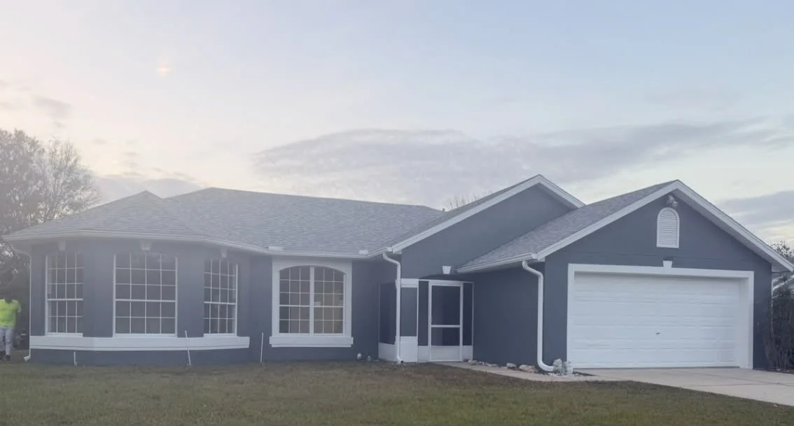 Front view of a blue single-story house with white trim, large windows, a two-car garage, and a front lawn with a person wearing a yellow shirt on the left side.