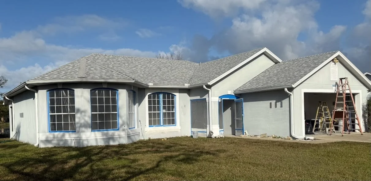 A house with a gray roof and white exterior undergoing exterior painting or renovation, with ladders and tools outside.