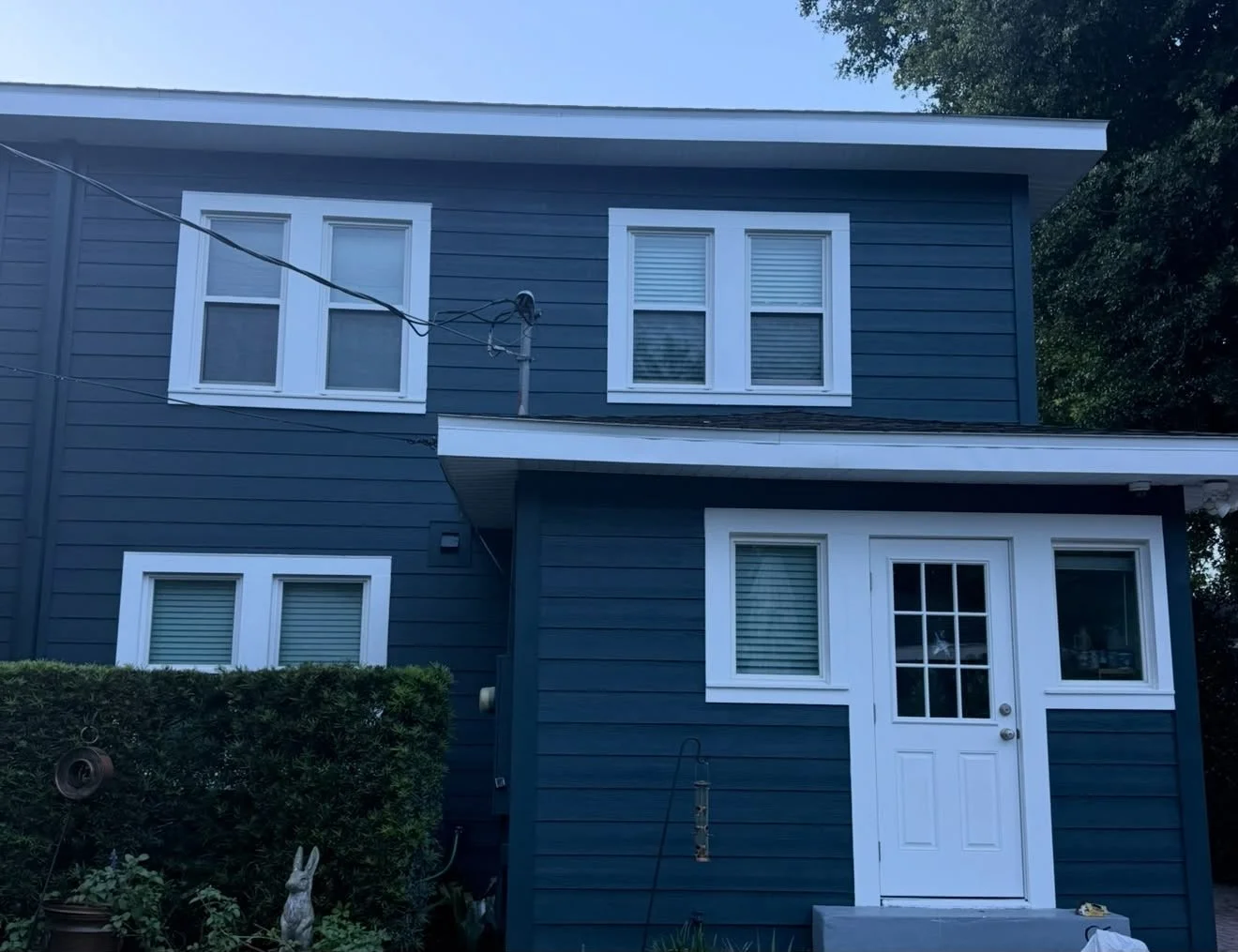 A two-story house painted dark blue with white window frames and a white door, surrounded by greenery and a hedge.