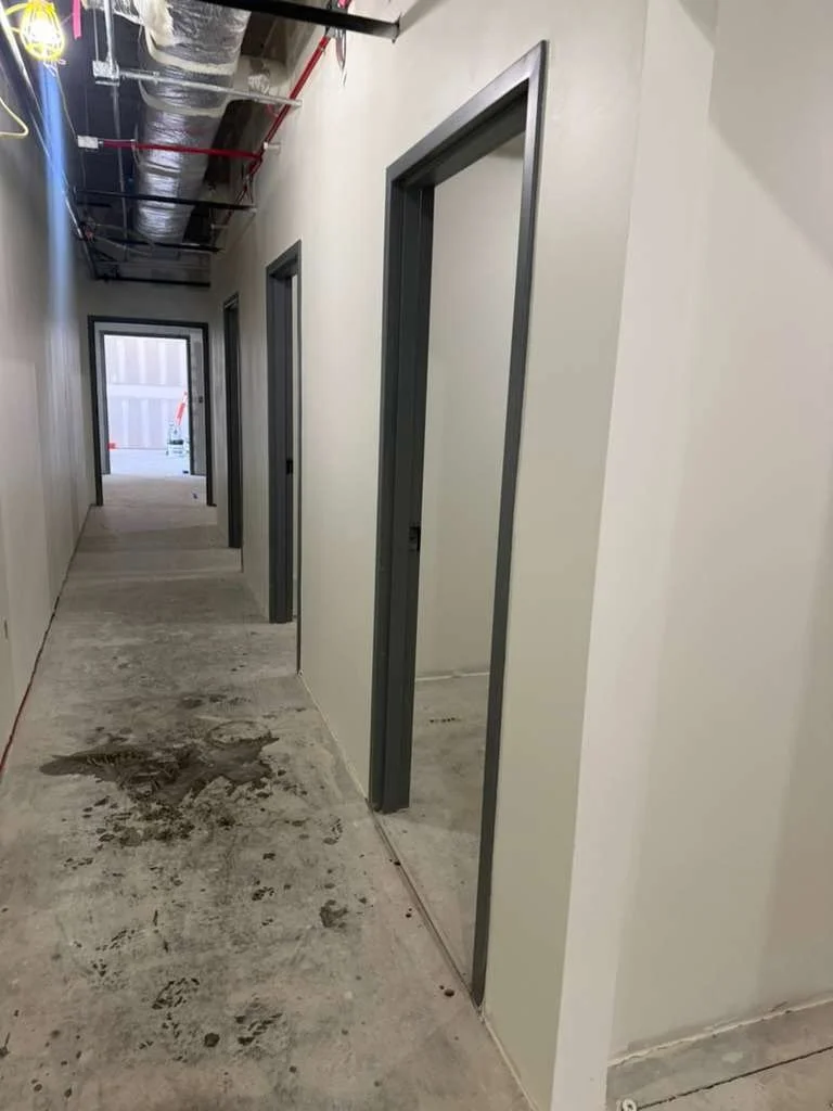 Construction hallway with three doorways on the right and a window at the end of the hall, exposed ceiling ducts, and concrete floor with dirt and footprints.