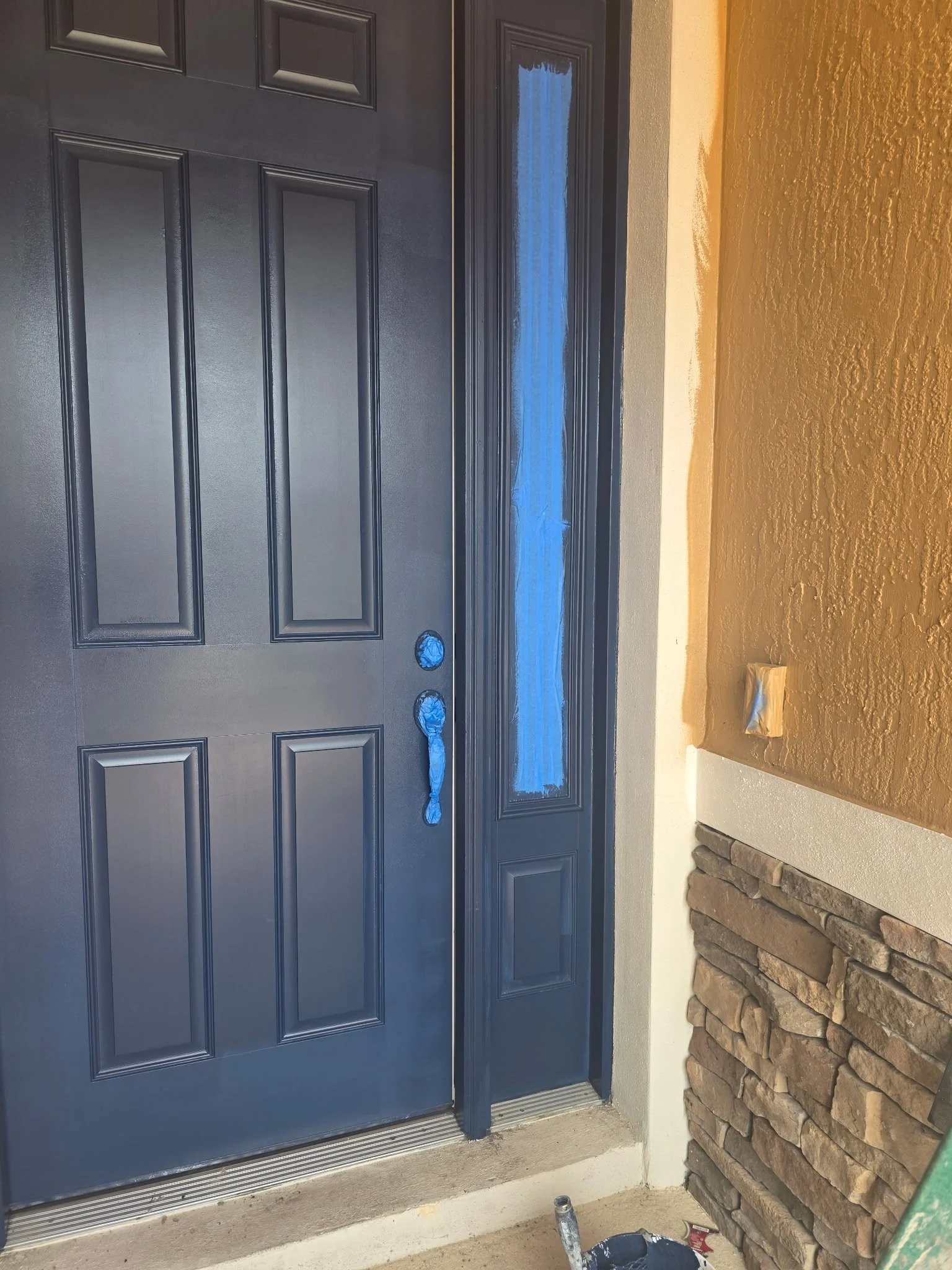 A black front door with small rectangular panels and a window at the top is surrounded by beige walls and a brick wall on the right side. Blue painter's tape covers parts of the door and window, indicating painting or renovation work. A small bucket and painting supplies are on the ground.