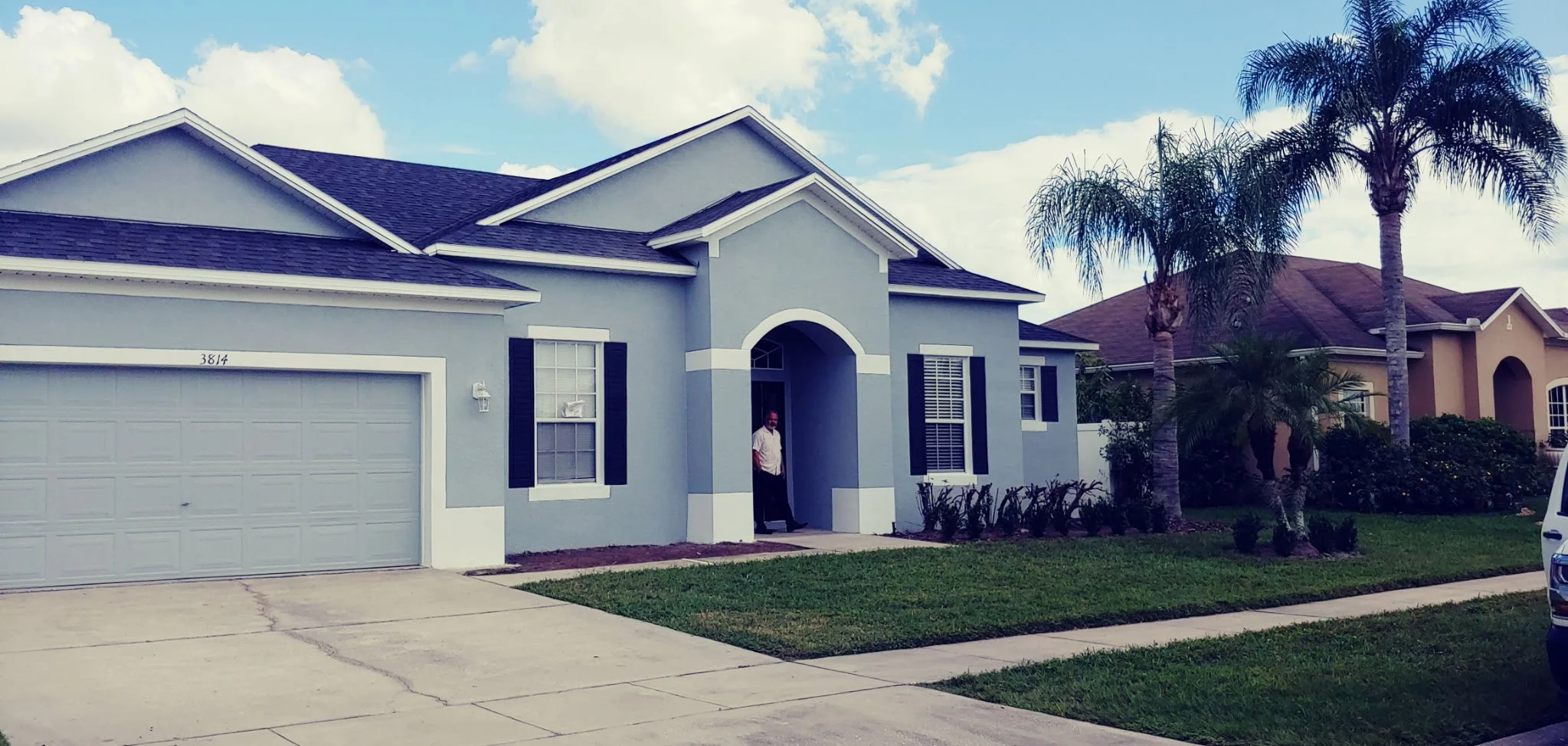 Light blue house with a white garage door, black shutters, and a man standing by the front door. The house has a small front lawn with grass, a concrete walkway, and tall palm trees in the yard. The sky is partly cloudy.
