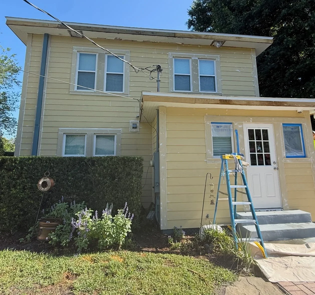 Two-story house with yellow siding, window frames taped for painting, a white back door, a blue ladder, and a small landscaped yard with plants and flowers, undergoing renovation or maintenance.
