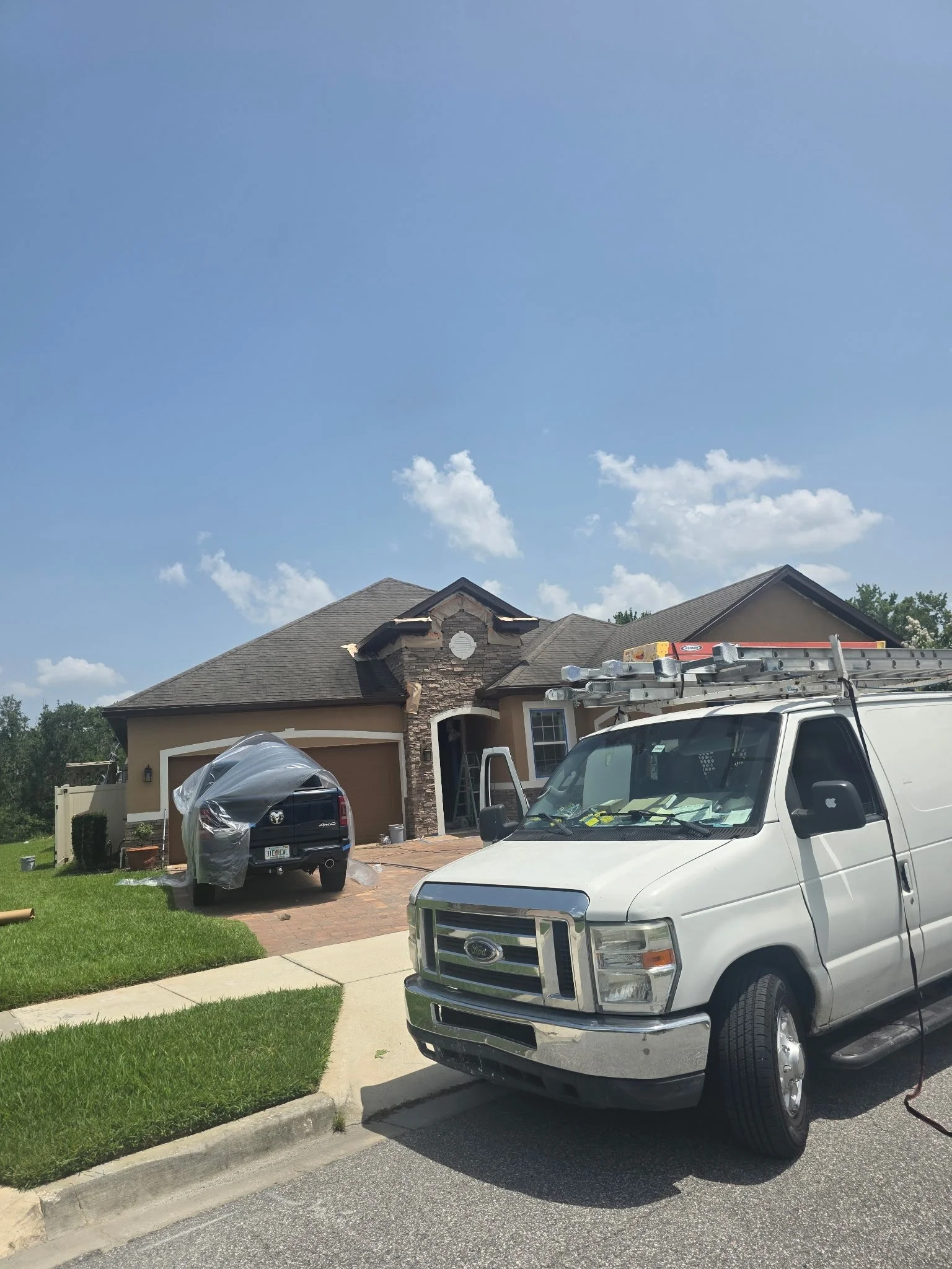 A house under renovation with construction trucks parked in the driveway. One truck has a ladder on top, and another is covered with plastic. The house has a stone facade with a gabled roof, and the sky is clear with a few clouds.