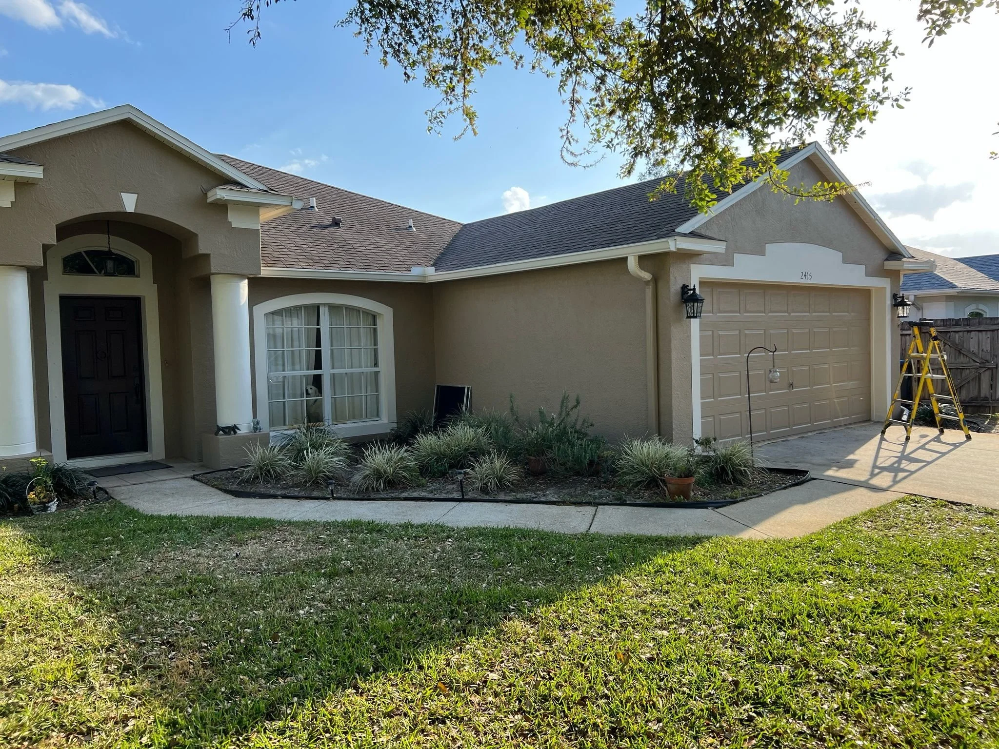 A beige house with a brown shingled roof, black front door, white columns, and a beige garage door with lanters on each side. There is a lawn in front with small plants and a sidewalk. A ladder is beside the garage.