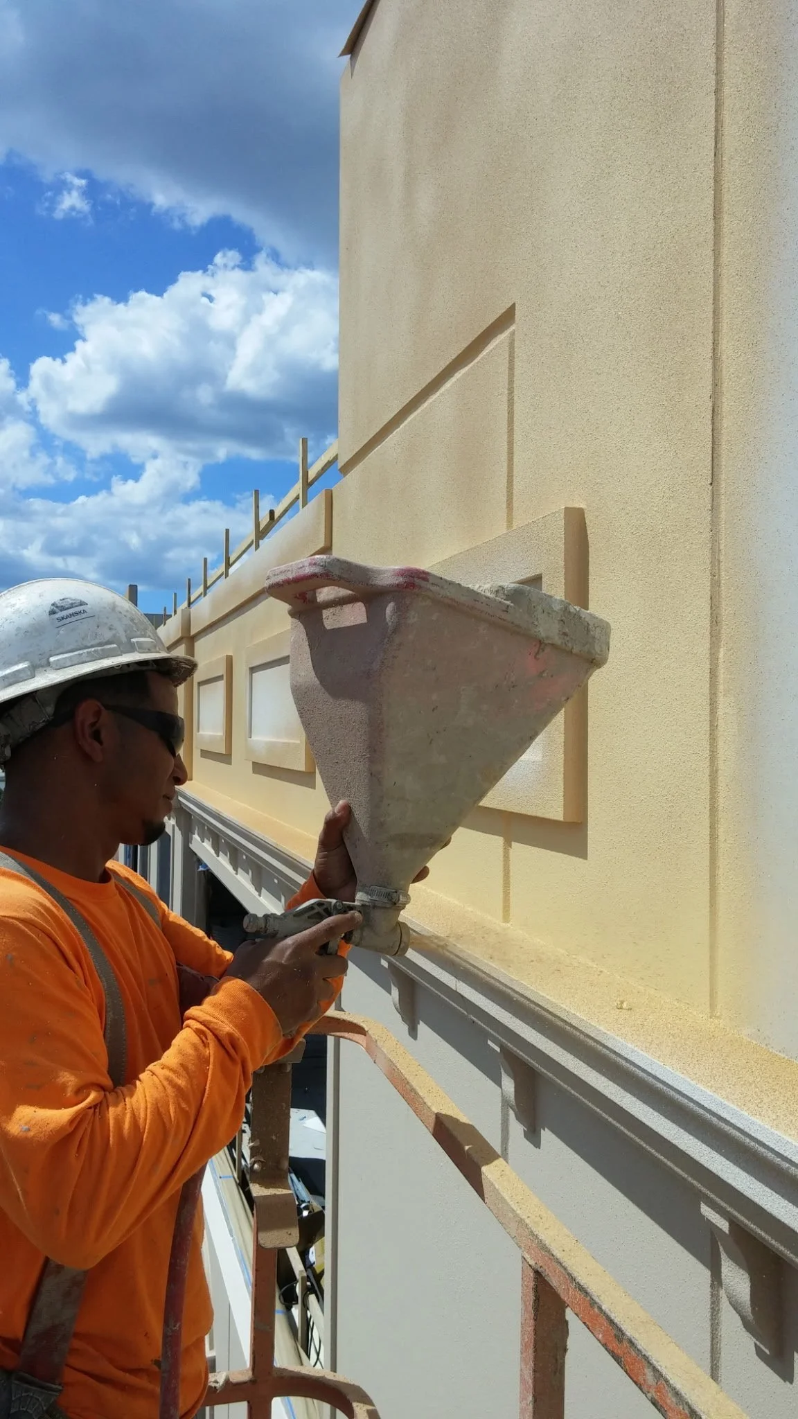 Construction worker wearing safety gear, pouring concrete onto a wall with a funnel and a handheld tool, with a building under construction and a blue sky with scattered clouds in the background.