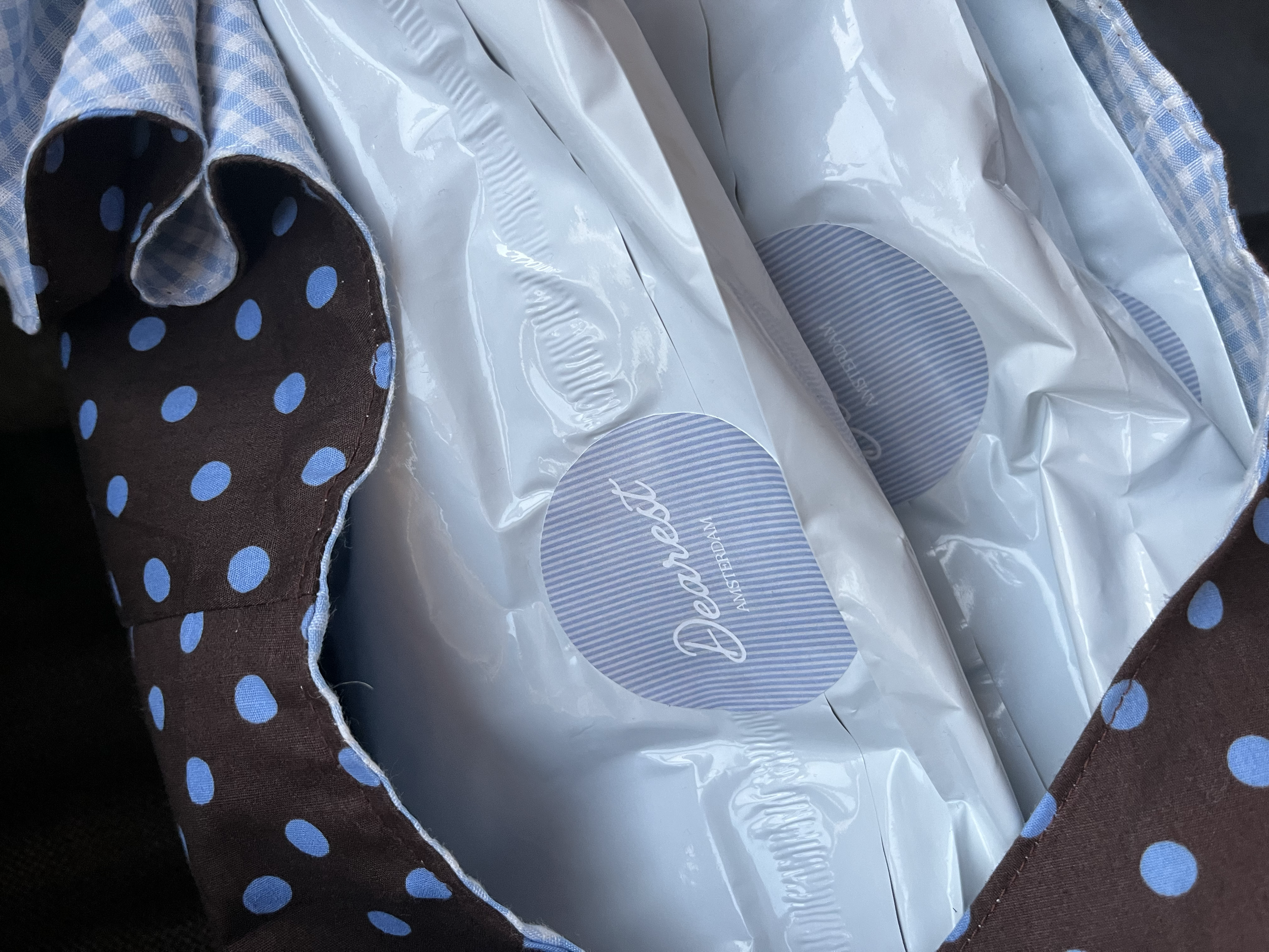 Close-up of a textured fabric with a checkered pattern in shades of blue and white, lining a bag. The bag contains a white plastic bag with blue stripes and a logo.
