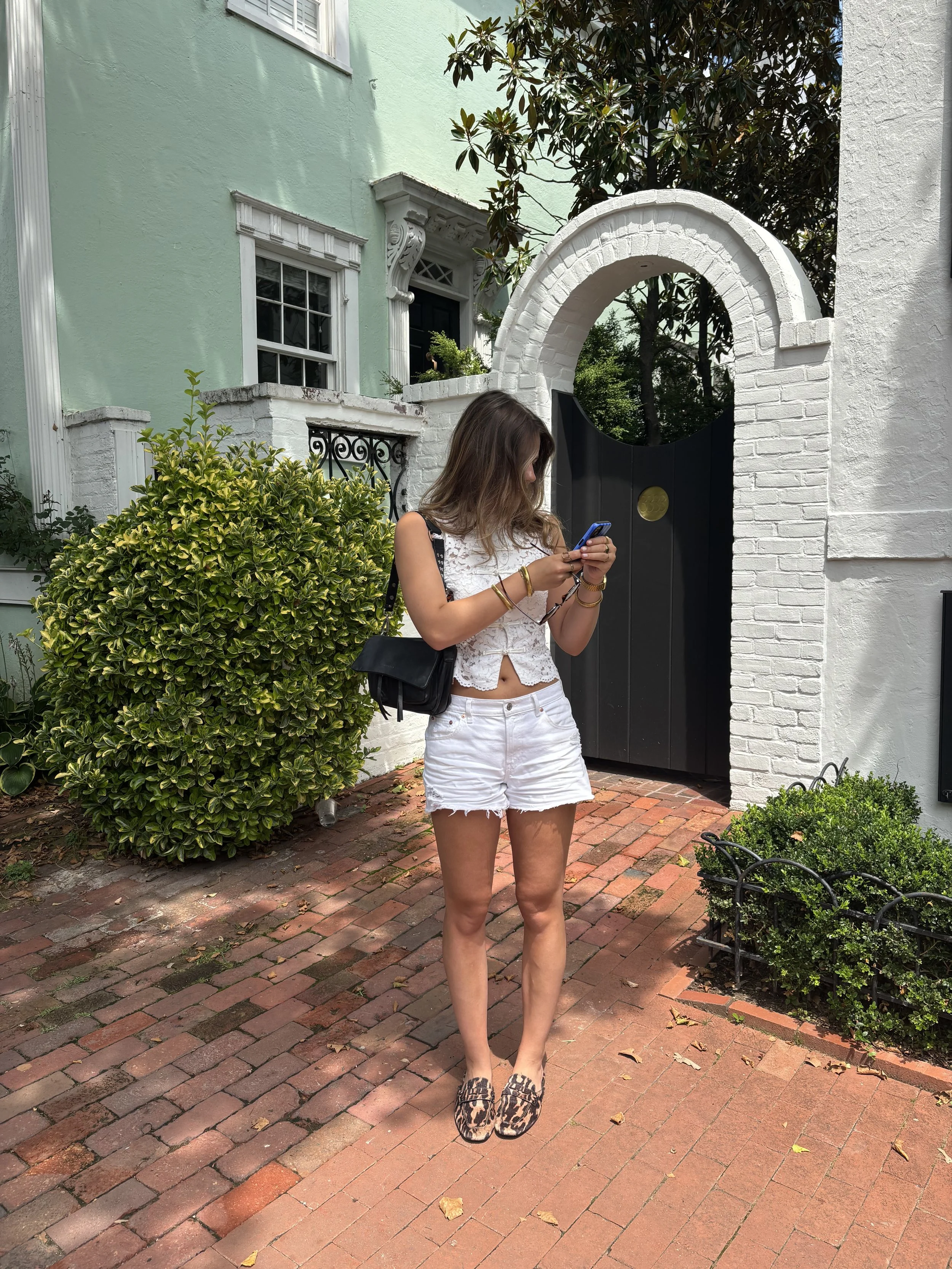 A young woman with long brown hair is standing outside on a brick pathway in front of a black gate with a white brick arch. She is looking at her phone and dressed in a white sleeveless lace top, white shorts, and leopard print slip-on shoes. She is wearing some gold bracelets and a black shoulder bag. There is a lush green bush to her left and a white-painted house with a large window and decorative trim in the background, along with a tree and some smaller plants.