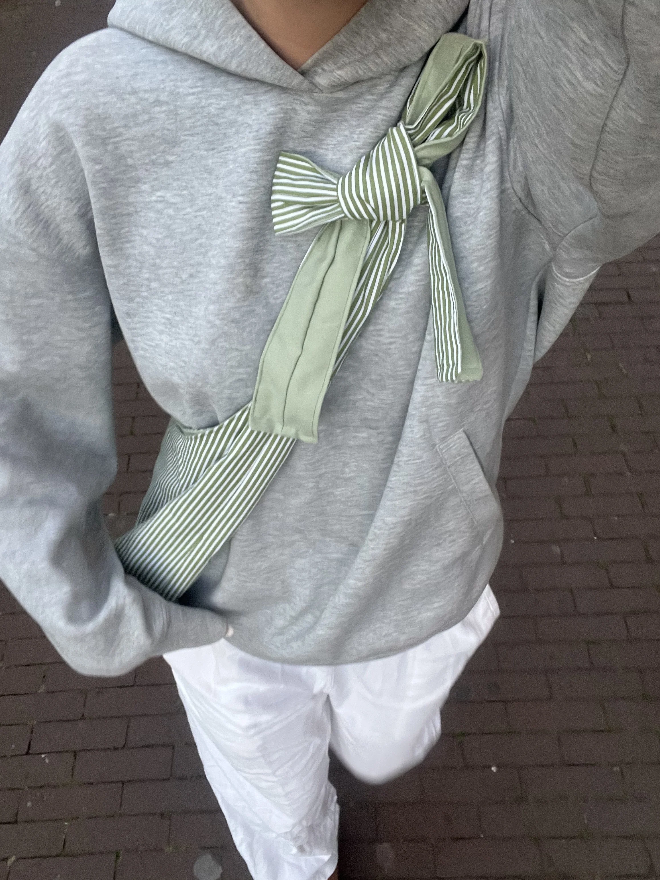 Person wearing a gray hoodie, white pants, and a green and white striped bow bag on their chest, standing on a brick sidewalk.