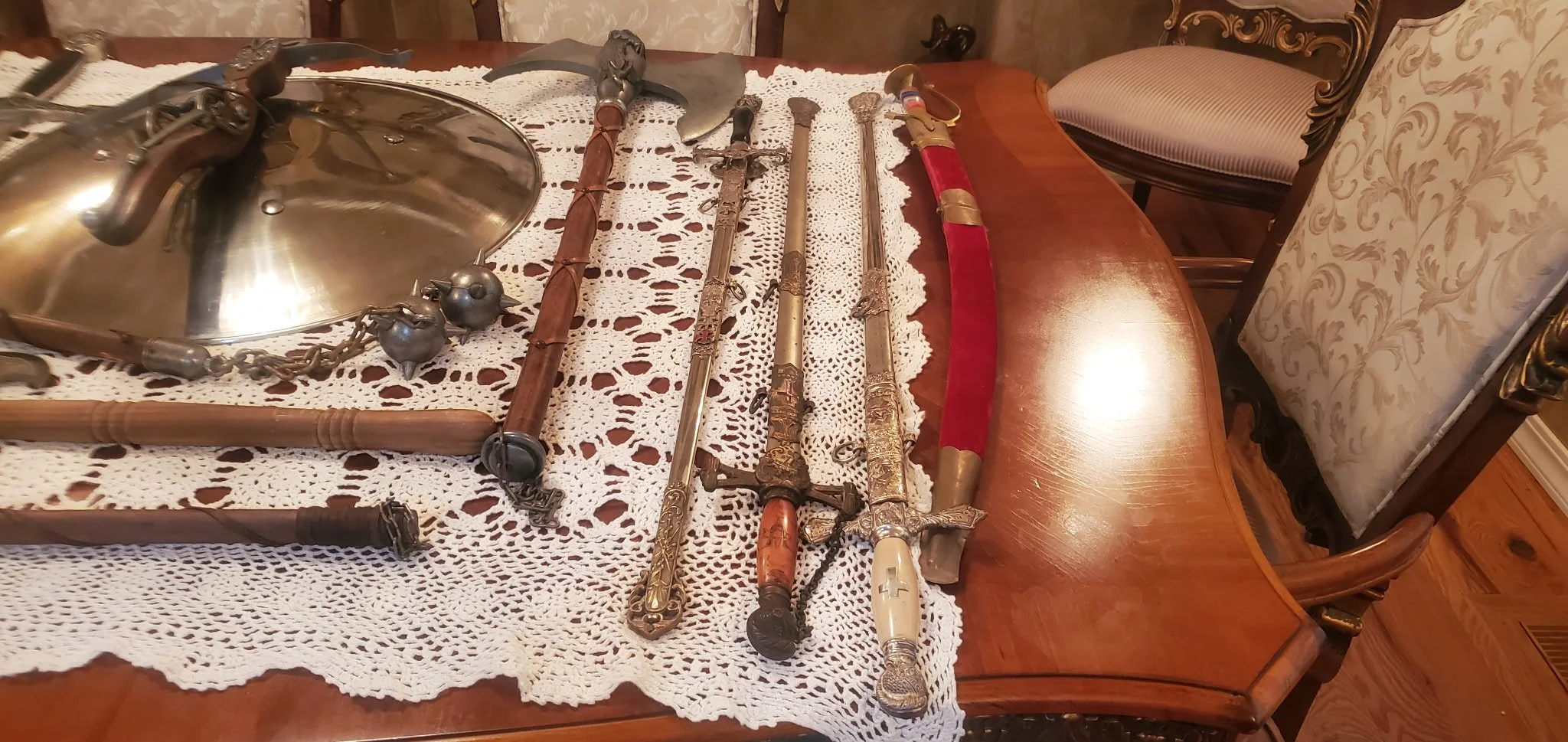 Collection of antique swords, knives, and a shield laid out on a lace tablecloth on a wooden dining table.