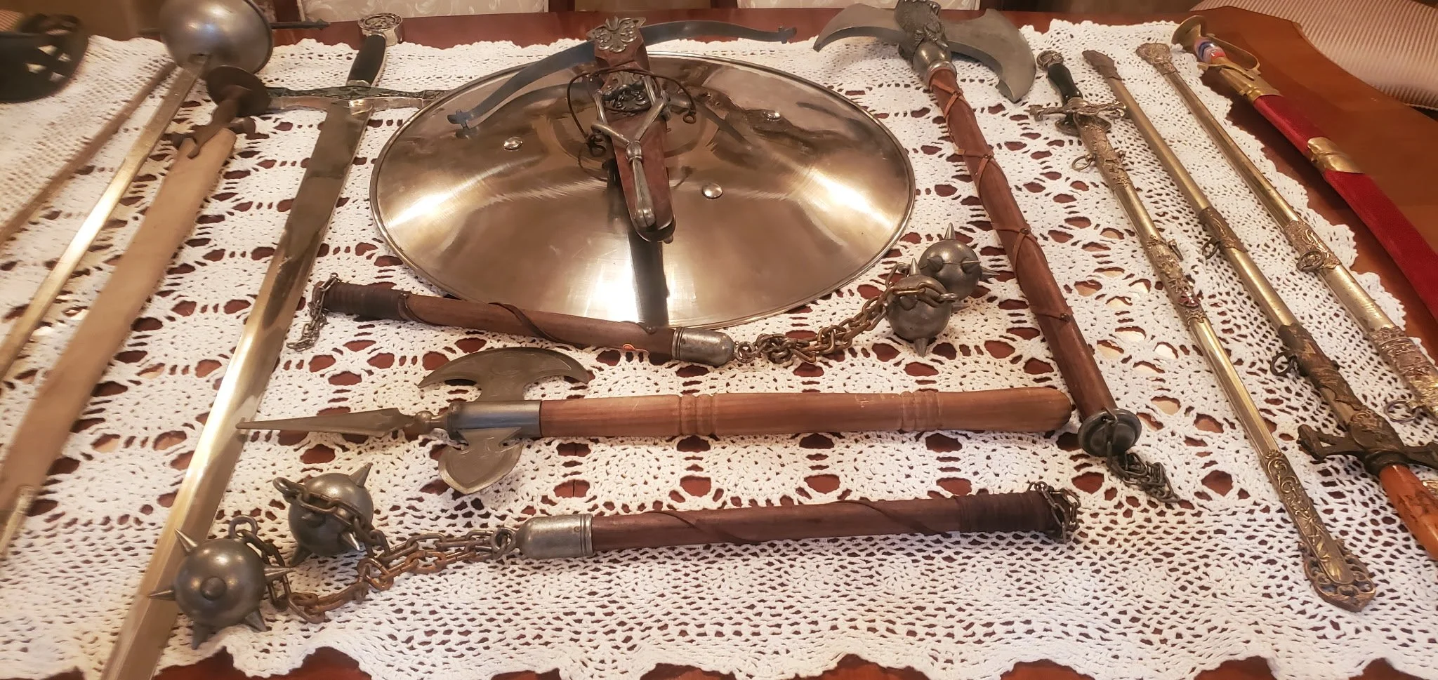 Collection of antique medieval weapons and armor, including swords, axes, maces, and a shield, displayed on a crocheted tablecloth.