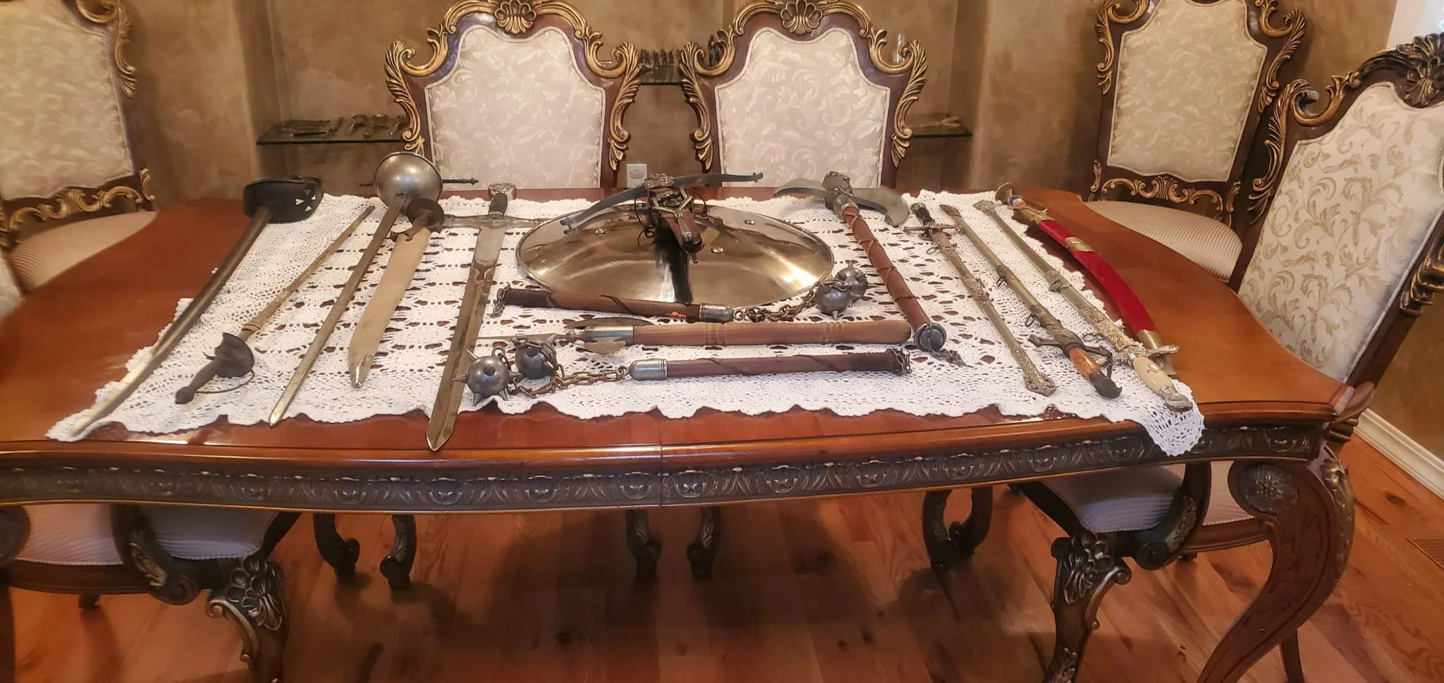 A wooden dining table with a lace tablecloth displays various antique swords, daggers, and a polished metal shield.