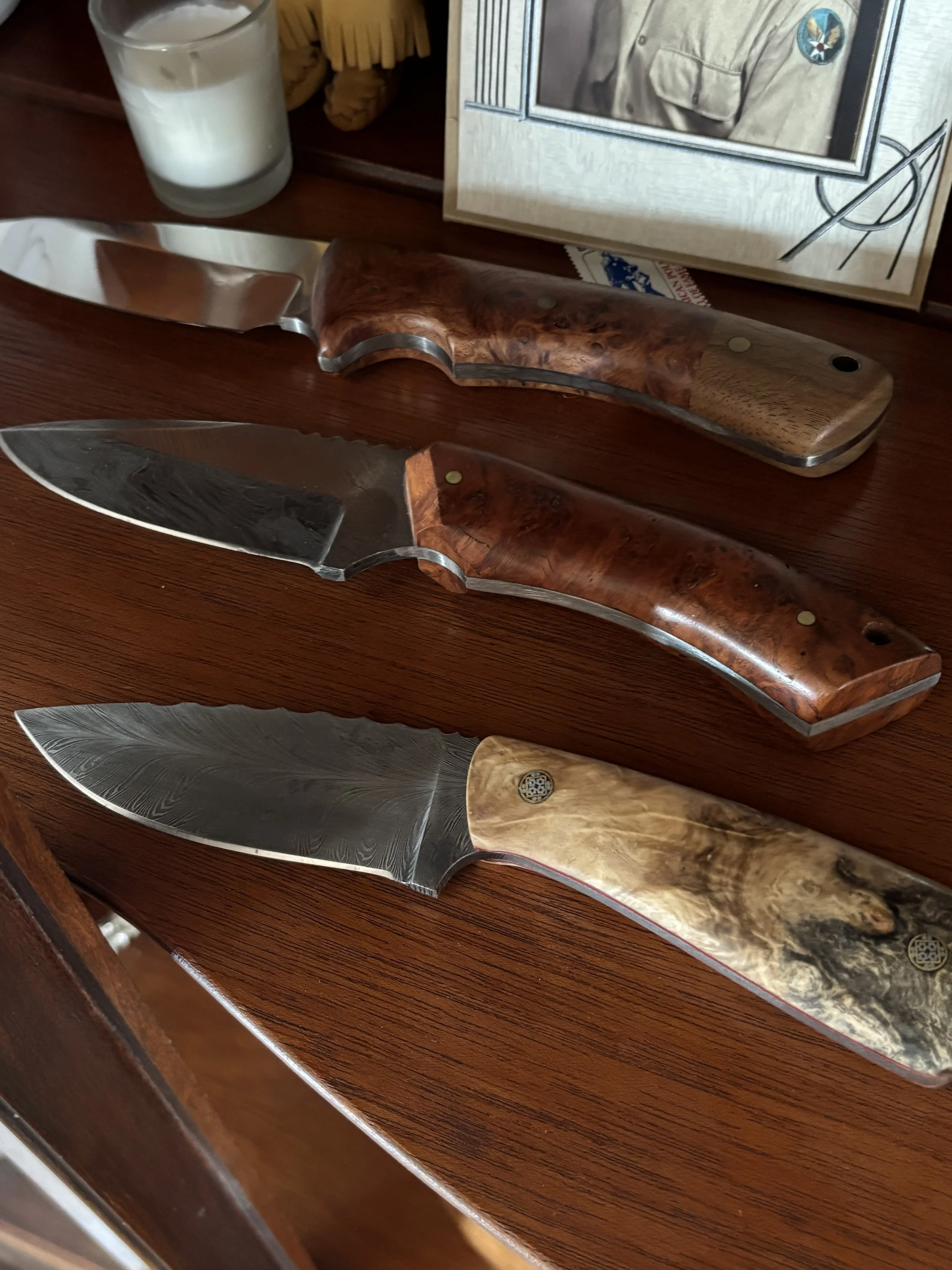 Three kitchen knives with ornate wooden handles placed on a polished wooden surface. A candle, a partially visible framed picture, and a magazine are also on the surface.