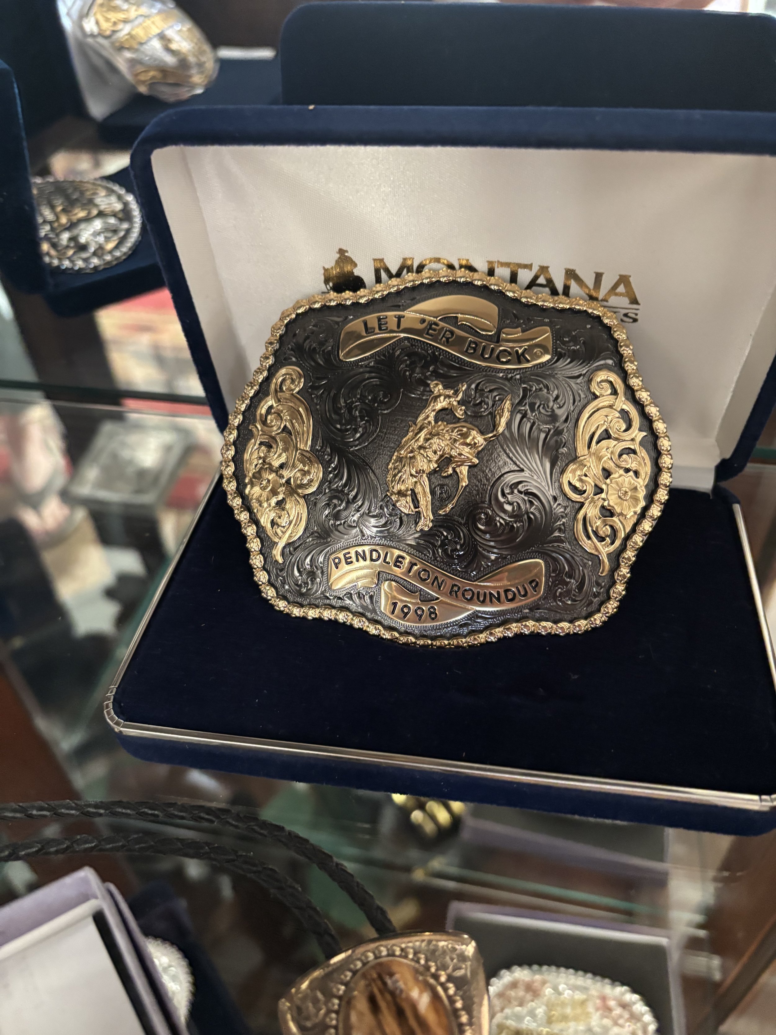 A decorative belt buckle featuring a detailed horse and rider with the words "Let 'er Buck" above and "Pendleton Roundup 1998" at the bottom, displayed in a jewelry box.