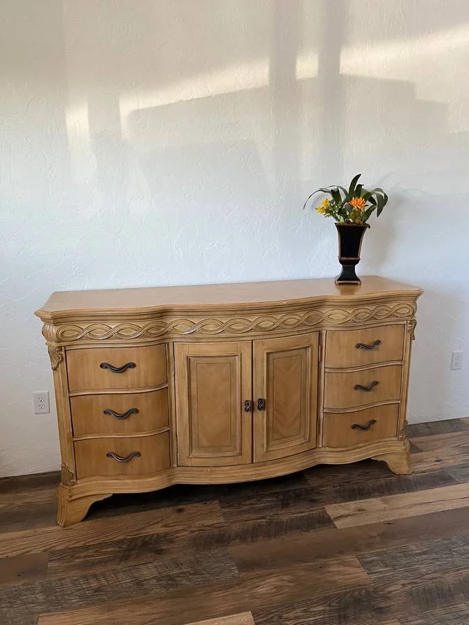 Wooden sideboard with decorative carvings and a flower vase on top, against a white wall with wood flooring.