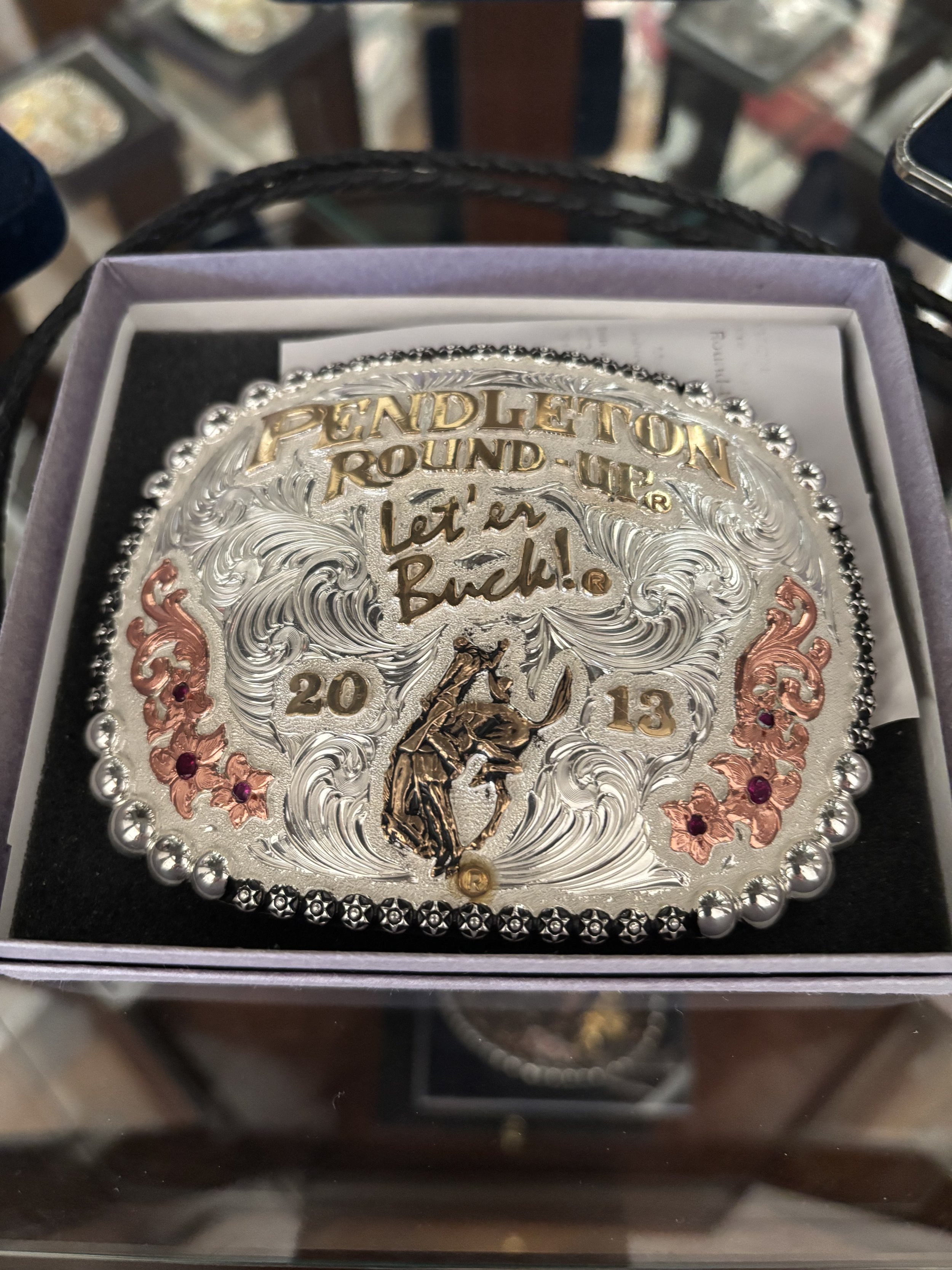 A decorative silver belt buckle with intricate swirling designs, gold lettering reading 'Pendleton Round-Up,' and pink accents. It is placed in a purple box, with a reflection of the photo surface visible below.