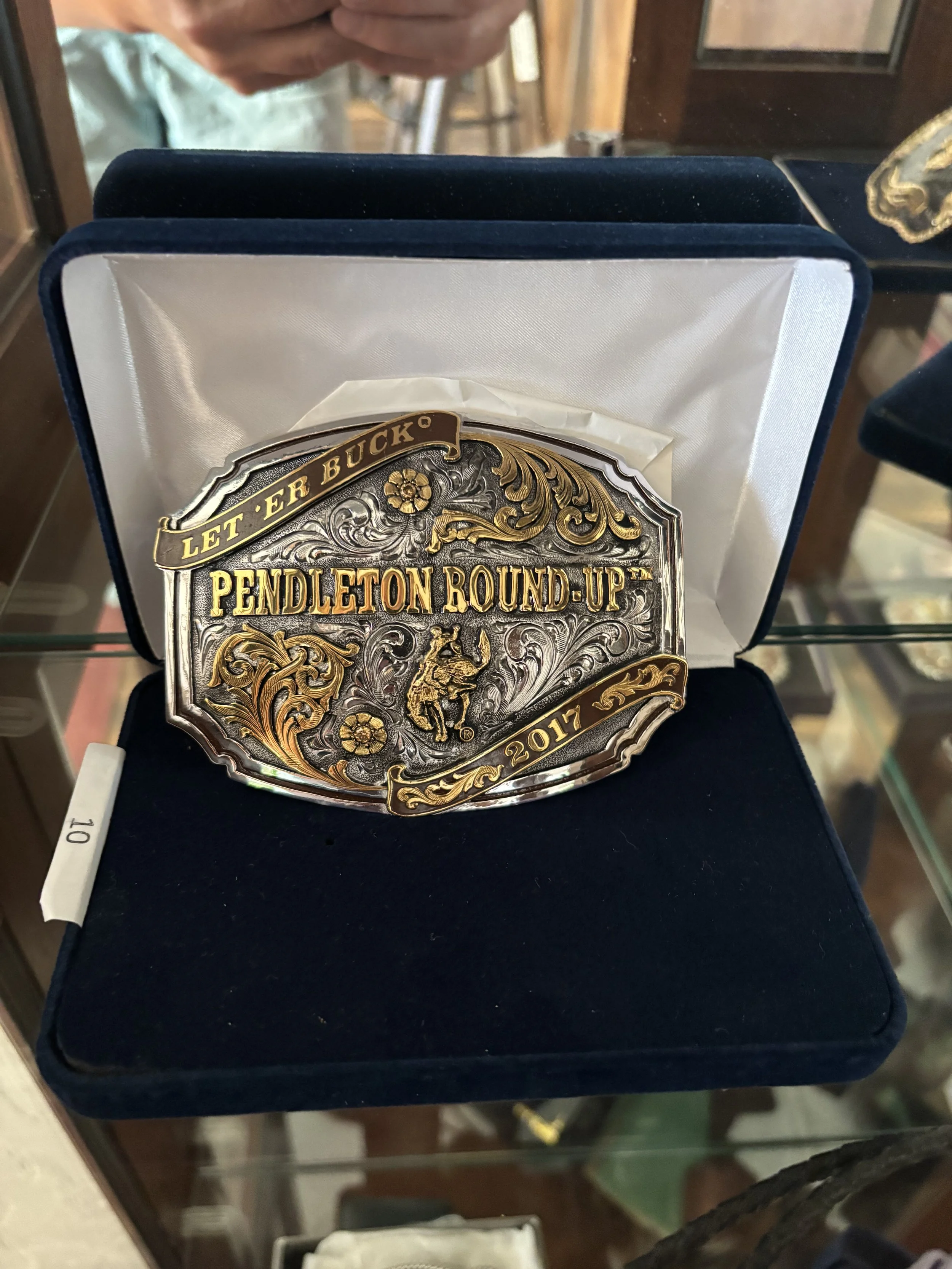 A belt buckle with ornate gold and silver design, inscribed with 'Let 'er Buck' and 'Pendleton Round-Up 2017', displayed in a black case.