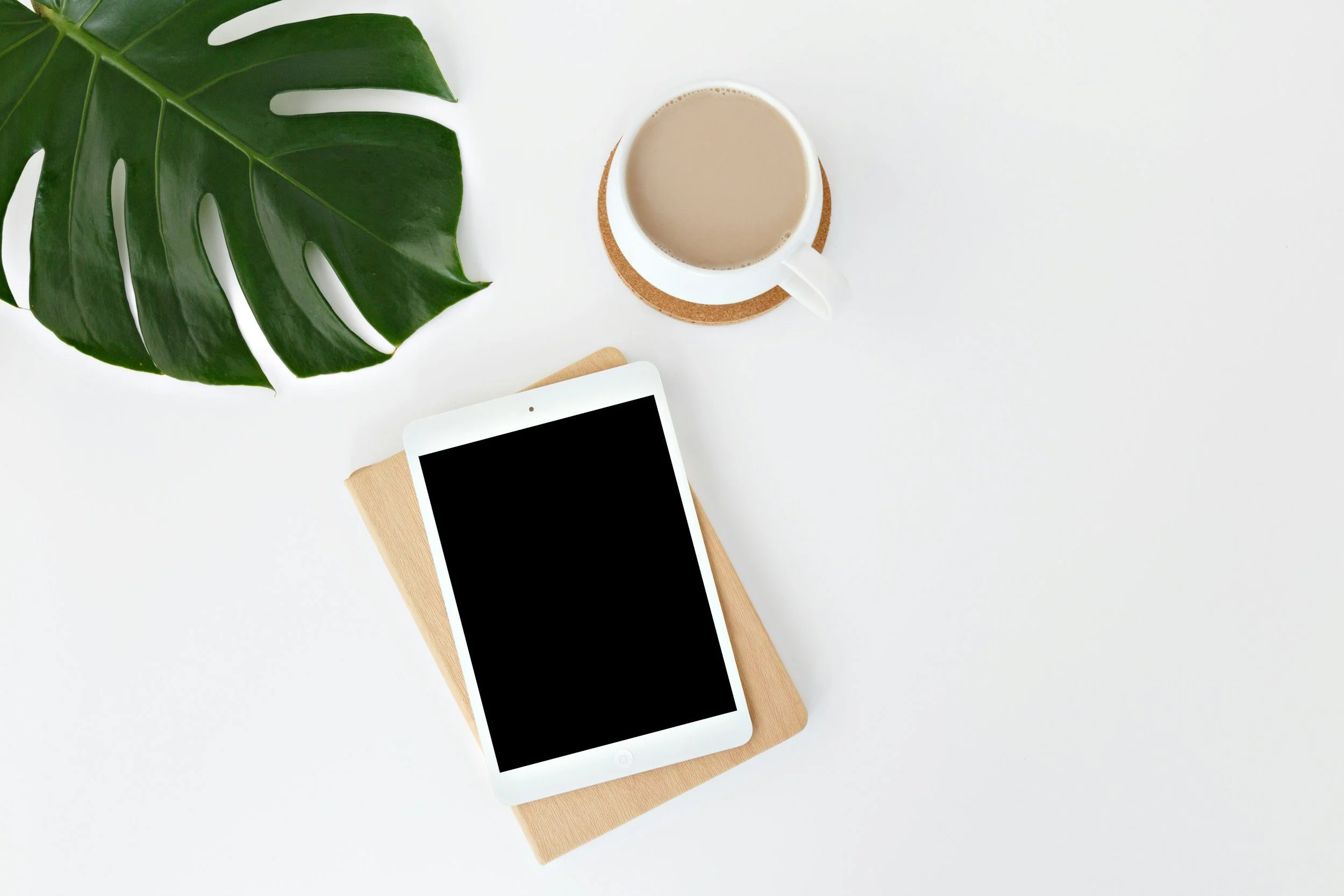 A white tablet with a black screen resting on a light wooden stand, next to a white mug filled with coffee or tea on a cork coaster, partially covered by a large green monstera leaf, on a white surface.