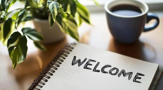 A spiral notebook with the word 'WELCOME' written on it, a potted plant with green leaves, and a cup of coffee on a wooden table.
