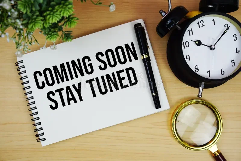Notebook with the words "Coming Soon Stay Tuned," a black pen, a gold magnifying glass, a black and white alarm clock, and a potted green plant on a wooden surface.