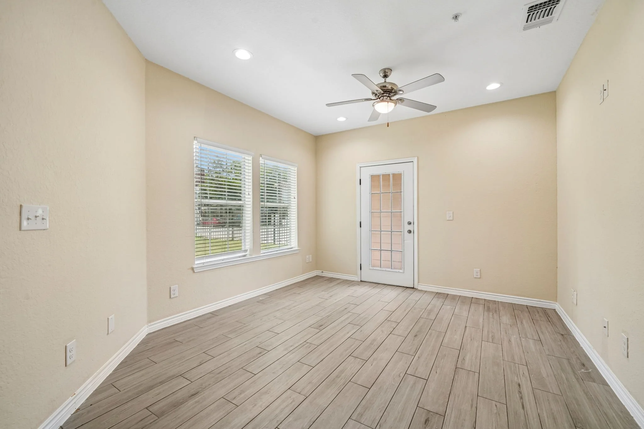 Empty living room with beige walls, hardwood flooring, two windows with blinds, and a glass-pane door leading outside. Ceiling fan with light. Recessed lighting in ceiling.