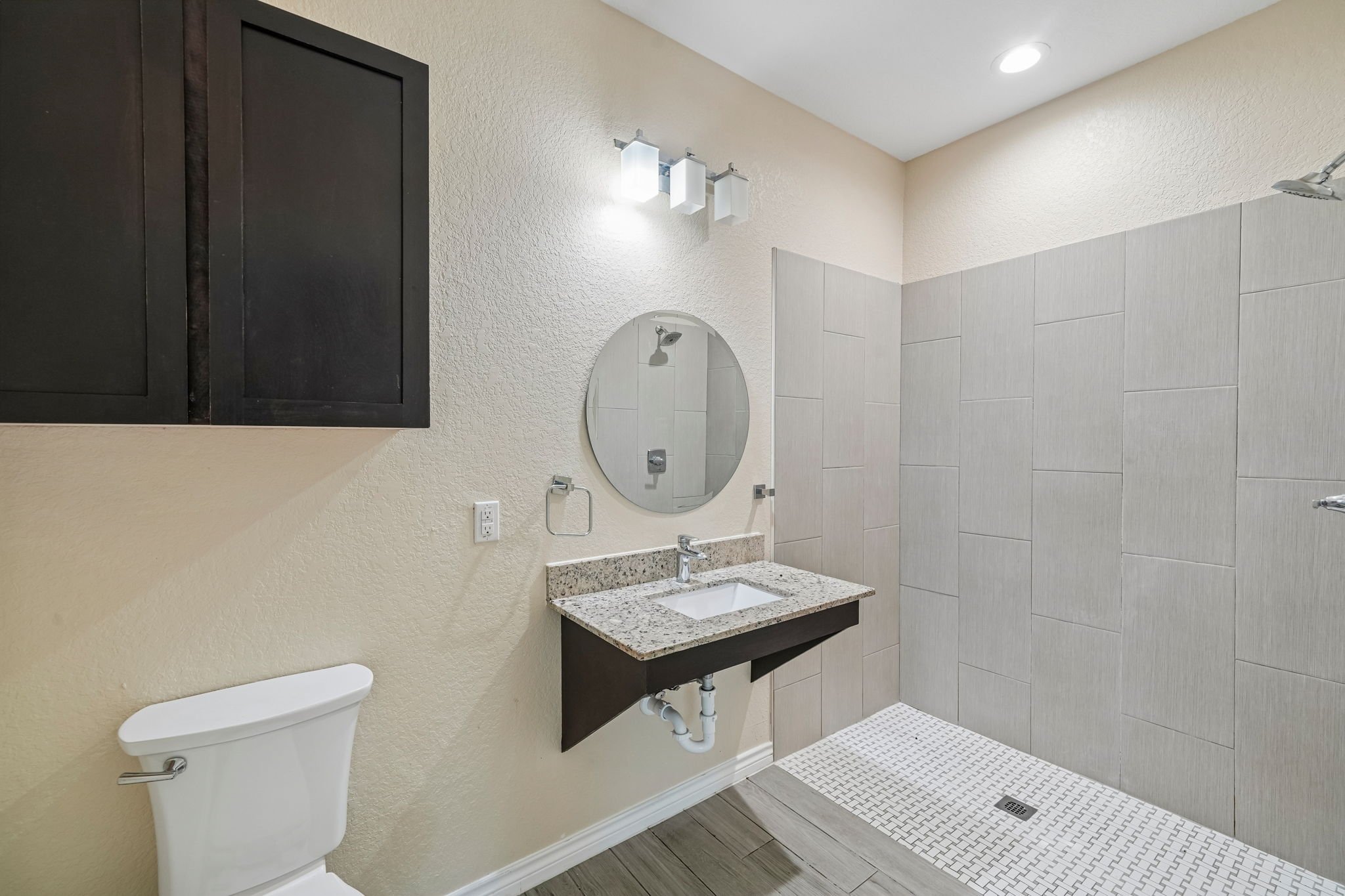 A bathroom with a beige textured wall, a black cabinet, a small oval mirror, a granite countertop with a built-in sink, a faucet, a white toilet, and a tiled shower area with white tiles, a shower head, a drain on the floor, and ceiling lights.
