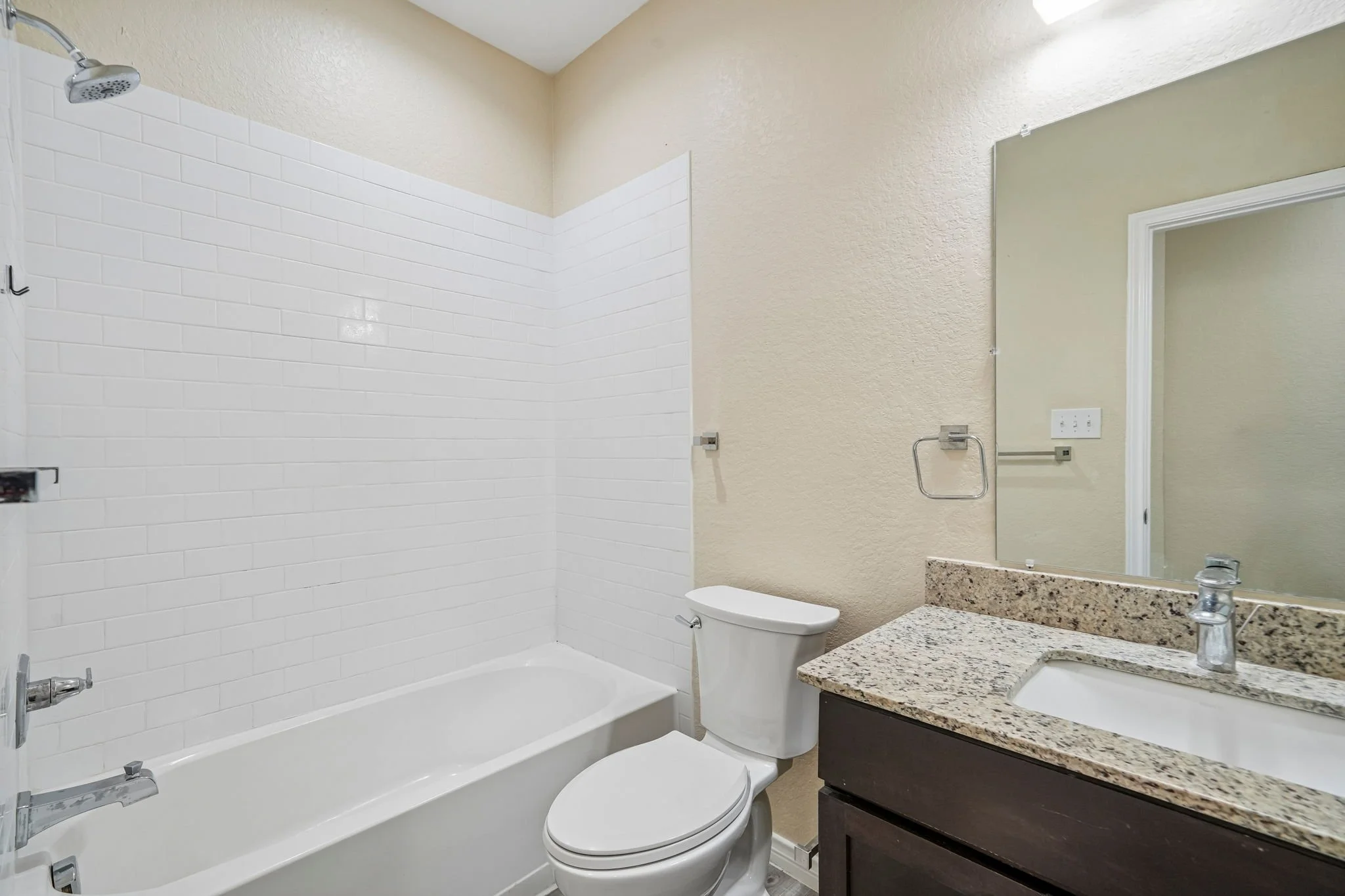 Bathroom with a bathtub with white tiles on the wall, a toilet, a granite countertop with a sink, a large mirror, a towel holder, and a window.