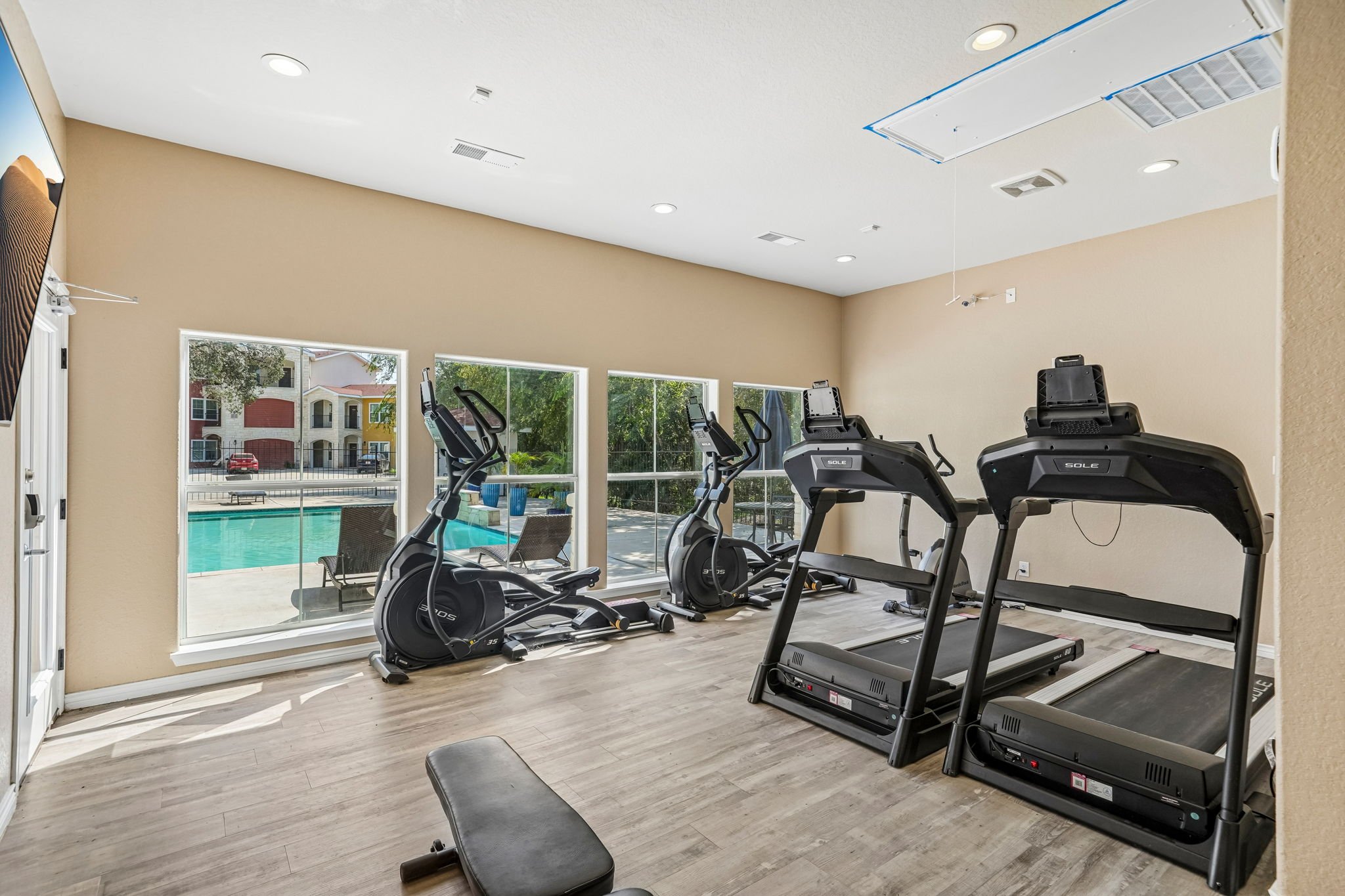 24-hour gym at La Marquesa Apartments located in the clubhouse with treadmills and elliptical machines near large windows showing outdoor pool and residential buildings.