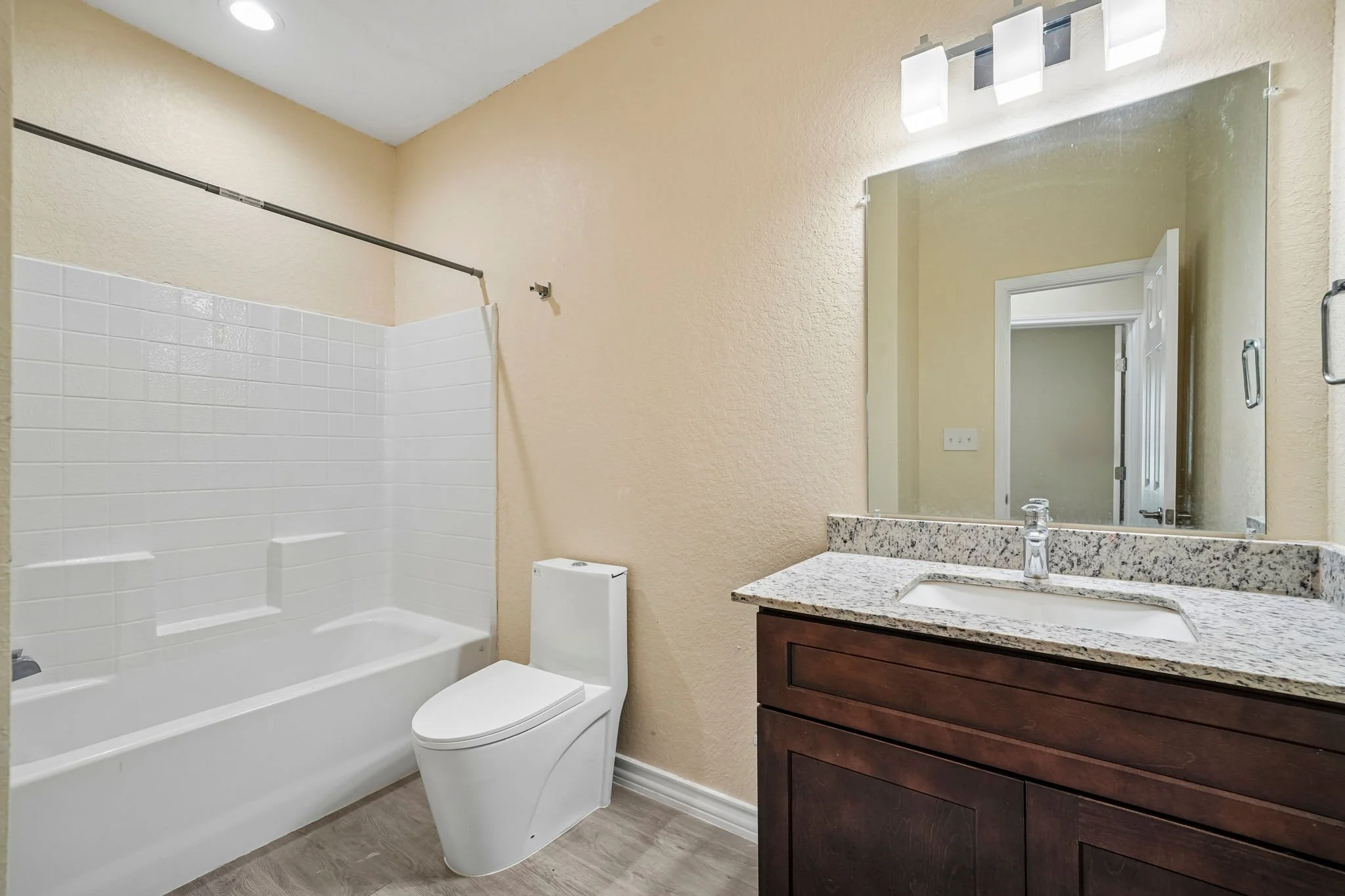 Bathroom with a bathtub and shower combination with a shower curtain rod, a toilet, a granite countertop with a sink, a mirror, and wall-mounted lighting fixtures.