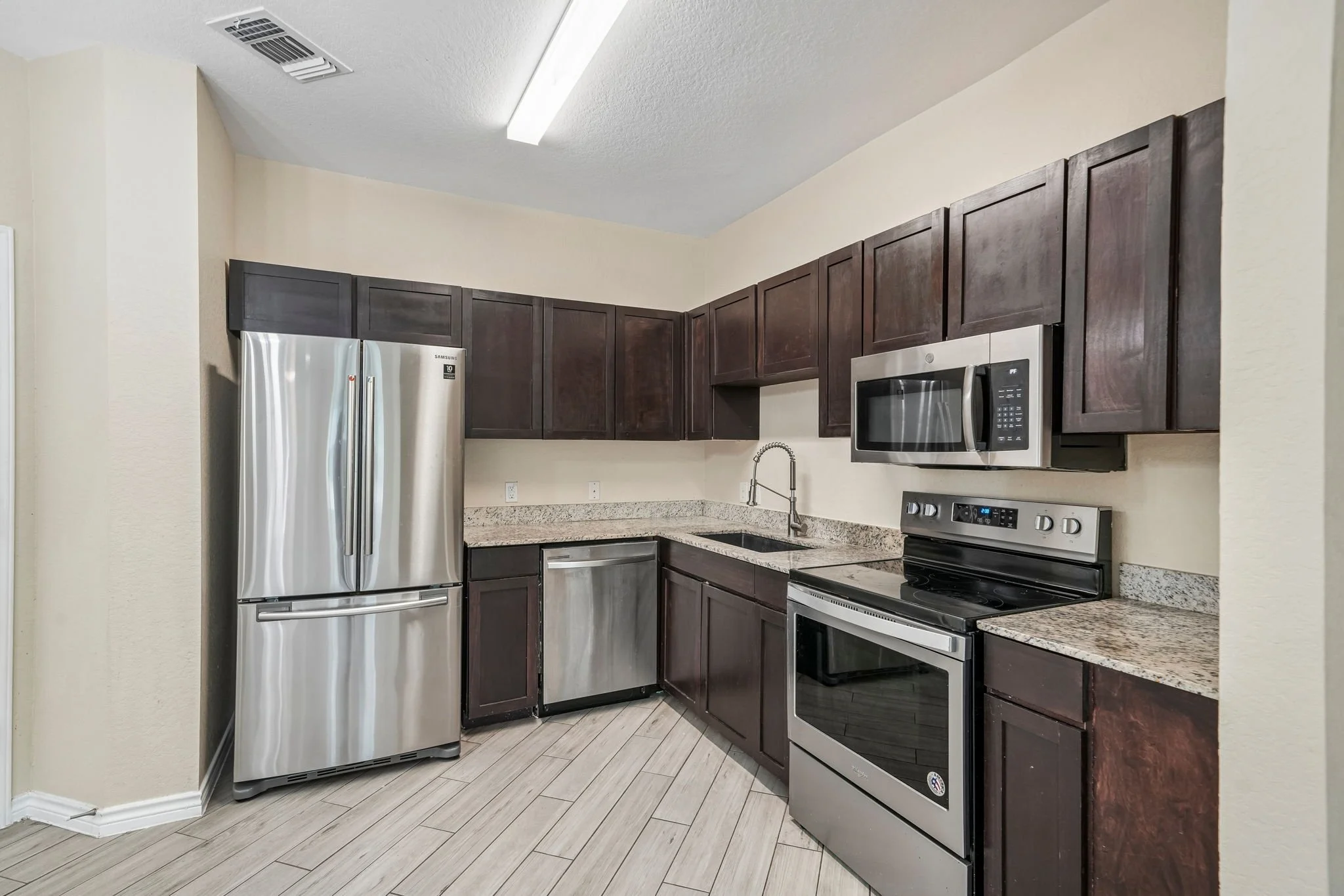 Kitchen with dark wood cabinets, stainless steel appliances including a refrigerator, microwave, and oven, granite countertops, and beige walls.