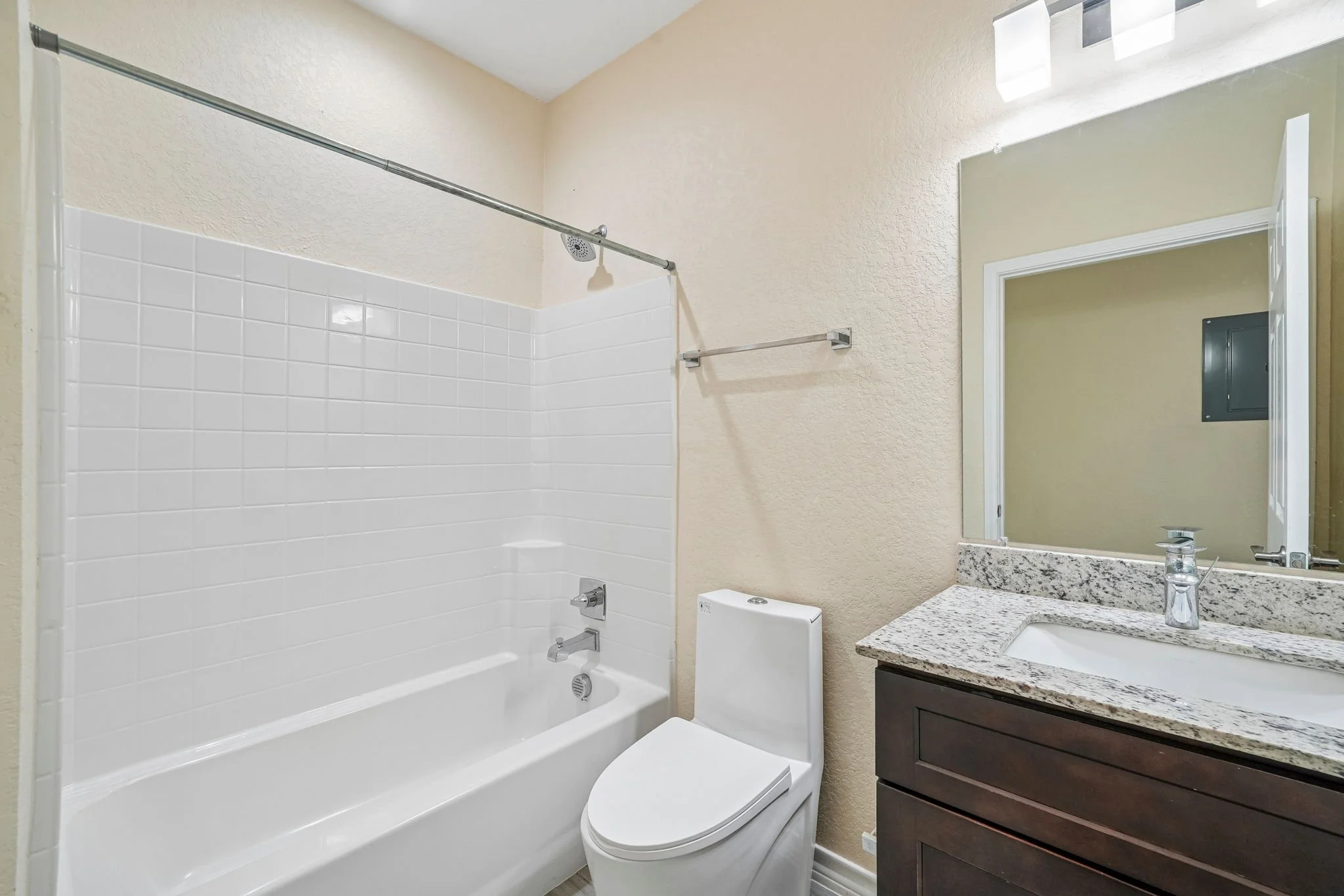 Bathroom with a white bathtub and shower area, a toilet, a granite countertop with a sink, mirror, and dark wood vanity, and beige textured walls.