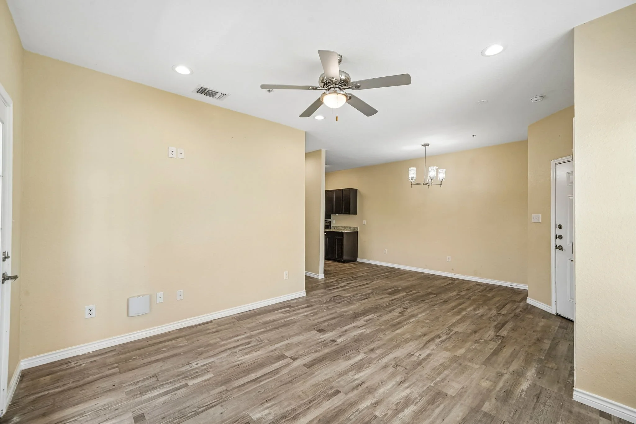 Empty living room with beige walls, wooden flooring, ceiling fan, mini chandelier, and kitchen in the background.