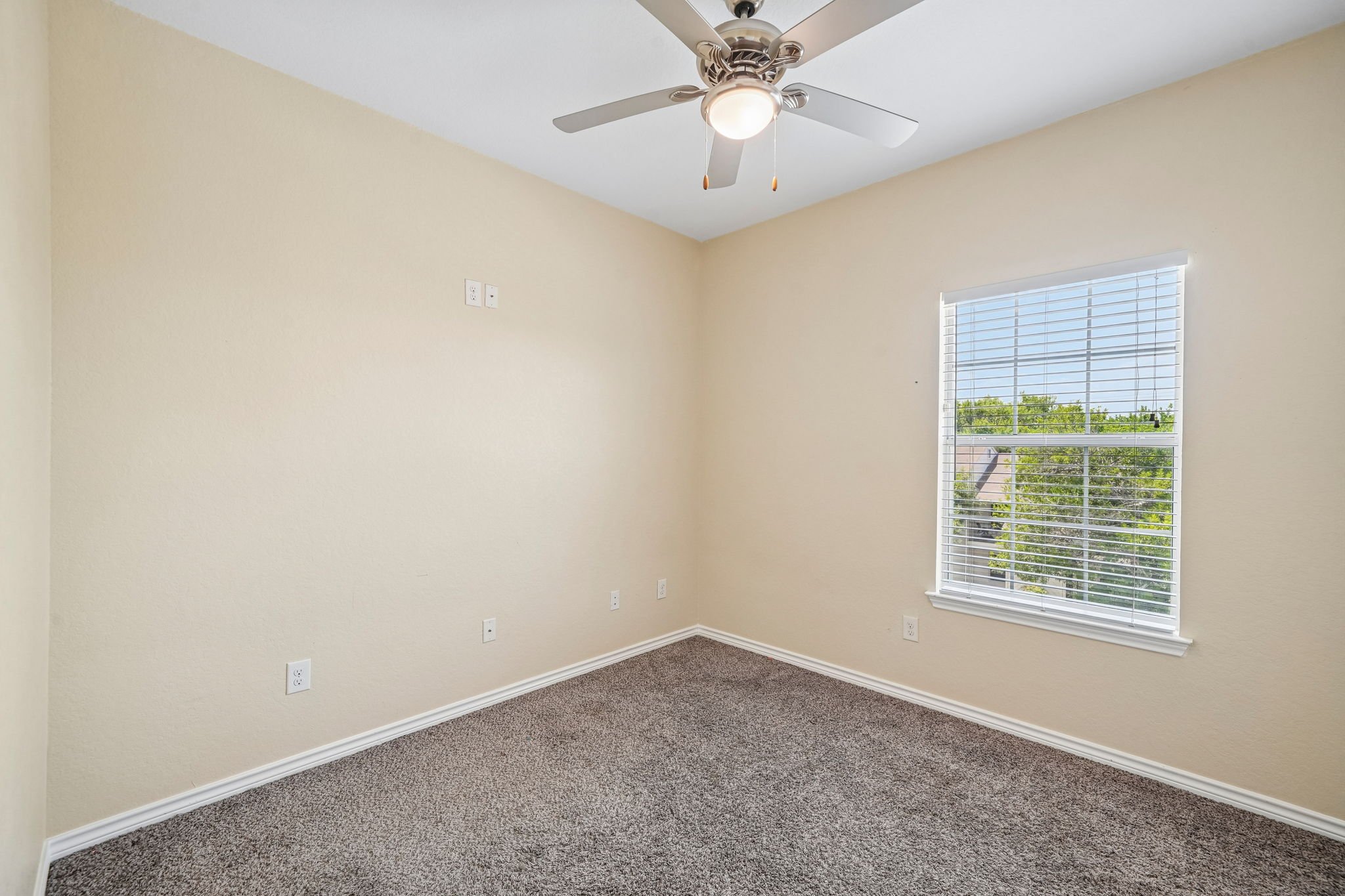 Empty bedroom with beige walls, a white ceiling fan, a window with blinds, and brown carpet flooring.