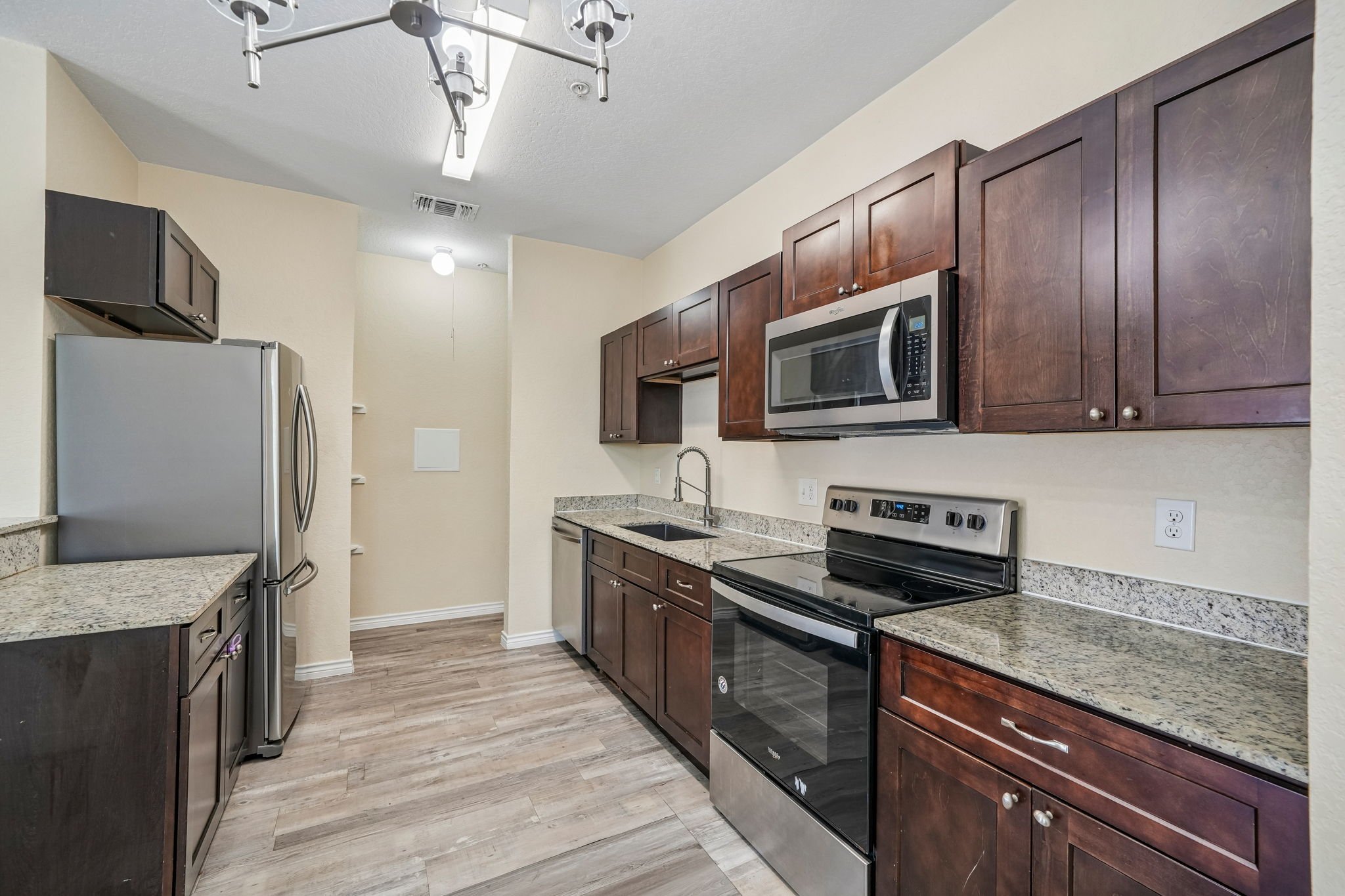 A modern kitchen with dark wooden cabinets, granite countertops, stainless steel appliances including a microwave and oven, and a refrigerator. The floor is wood-like laminate, and there are ceiling lights.