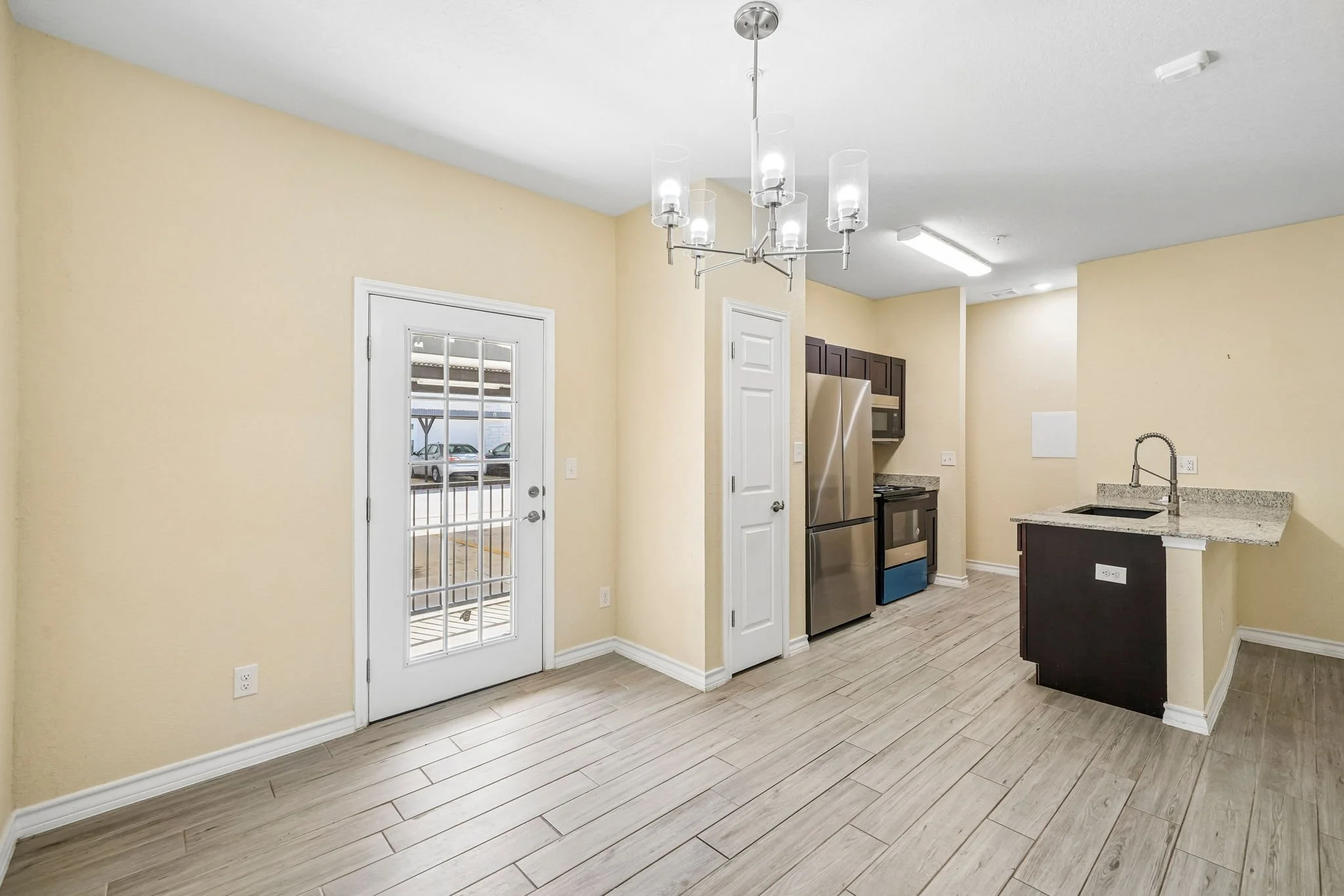 Interior view of a modern kitchen with light-colored walls, wood-look flooring, stainless steel refrigerator, microwave, and oven, a small kitchen counter with a sink, and a glass-paneled door leading outside.