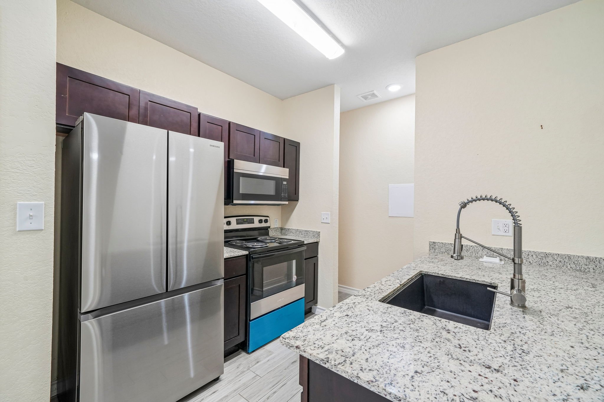 A modern kitchen with stainless steel refrigerator, microwave, and stove. Dark wood cabinets, granite countertops, a black sink, and a gooseneck faucet. Bright lighting with a white ceiling and light-colored walls.