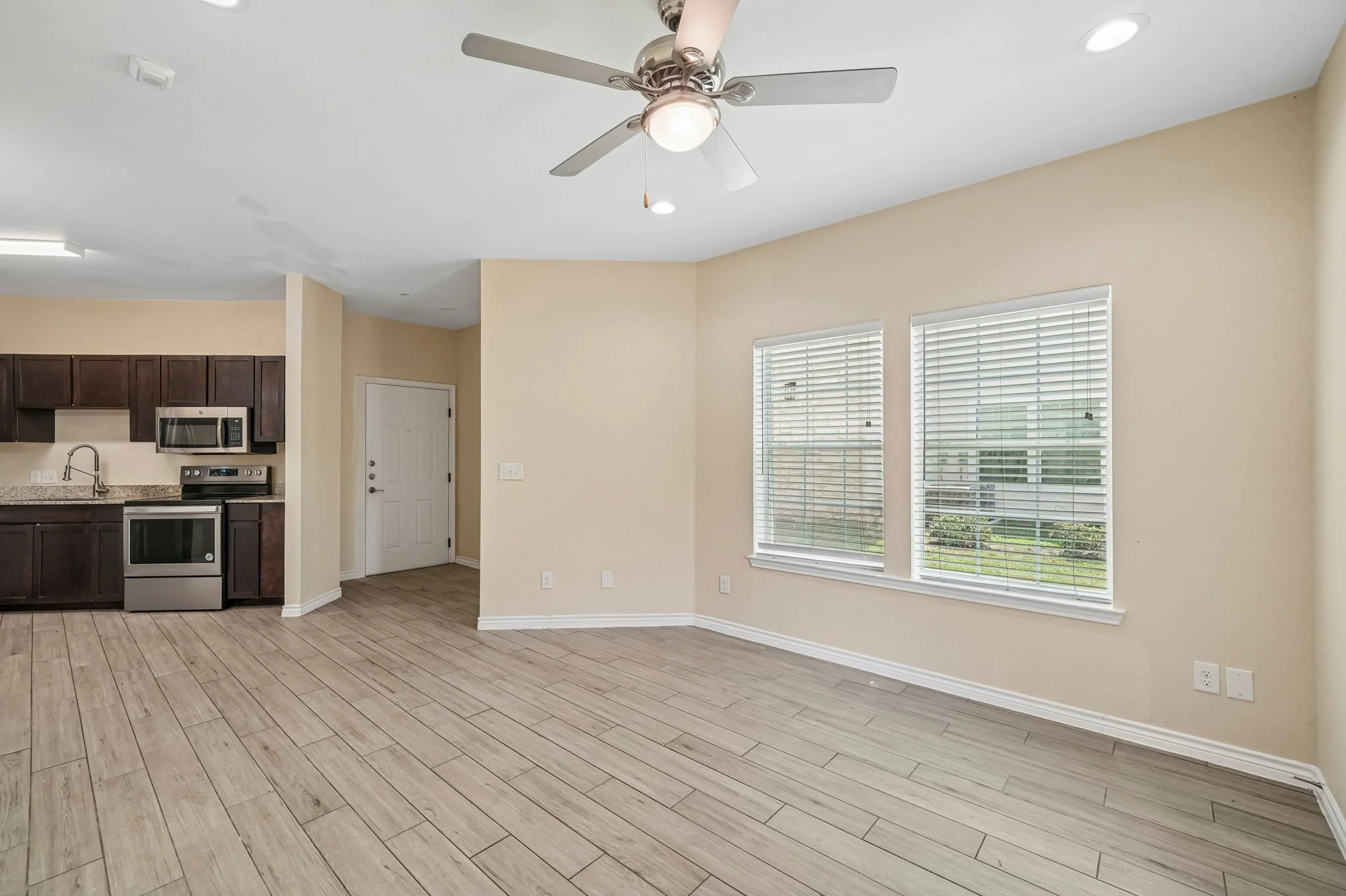 Bright living space with large windows, beige walls, wood-like flooring, ceiling fan, and open kitchen area with dark cabinets and stainless steel appliances.
