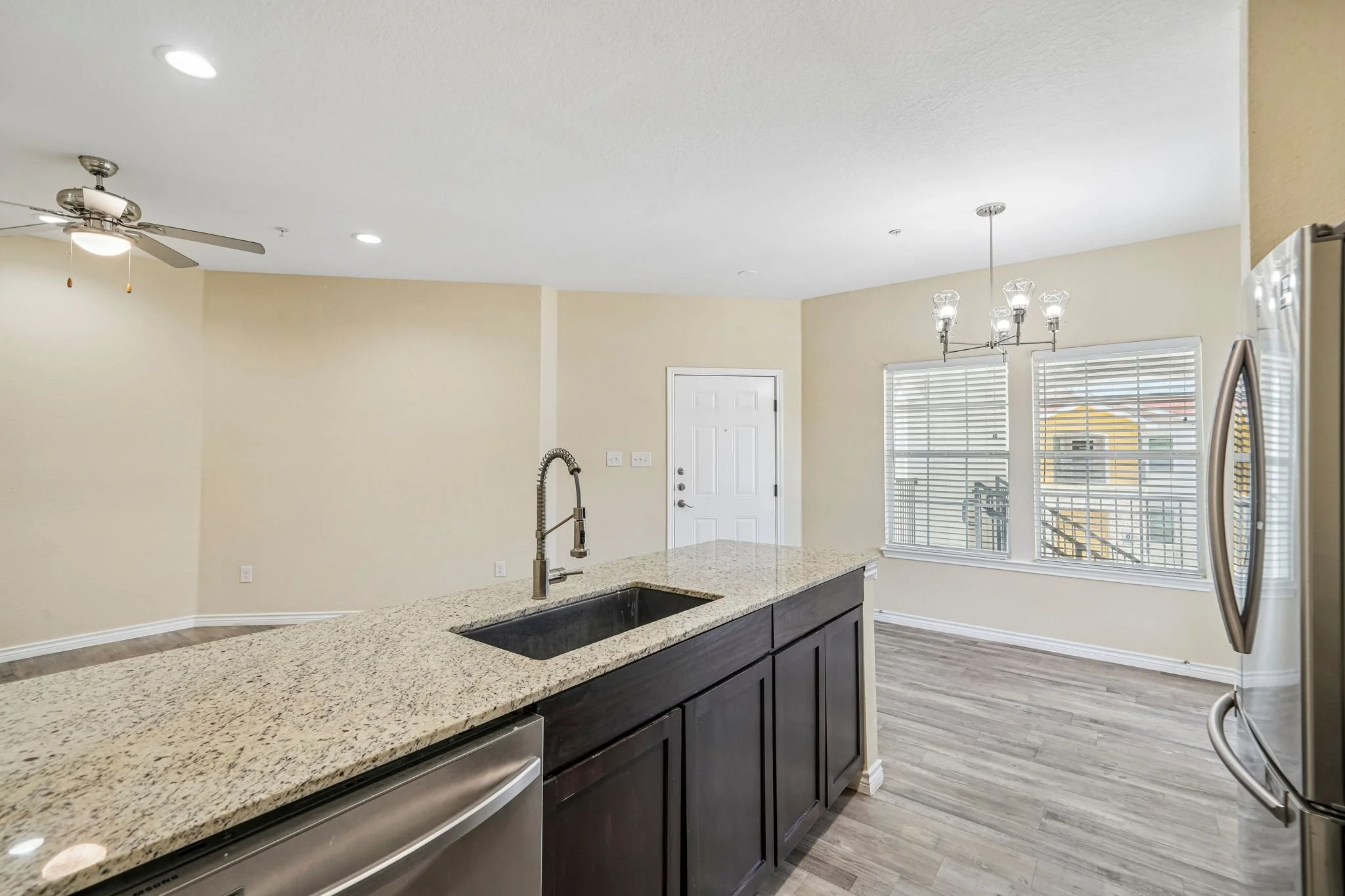 Modern kitchen with a granite countertop, dark cabinets, a stainless steel dishwasher, a sink with a tall faucet, and large windows with blinds. Recessed lighting and a hanging light fixture are visible, along with a ceiling fan.