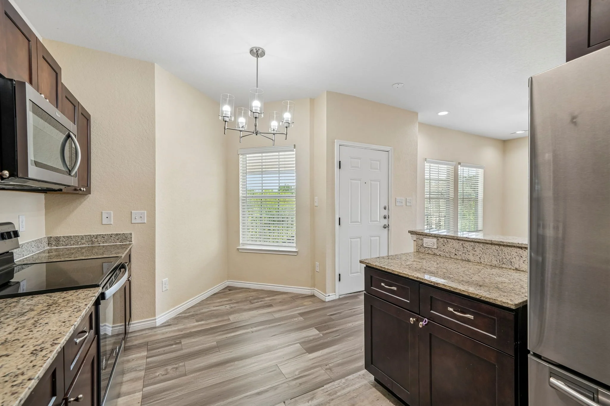 Modern kitchen with granite countertops, dark wood cabinets, stainless steel appliances, and large windows with blinds, illuminated by a contemporary chandelier.