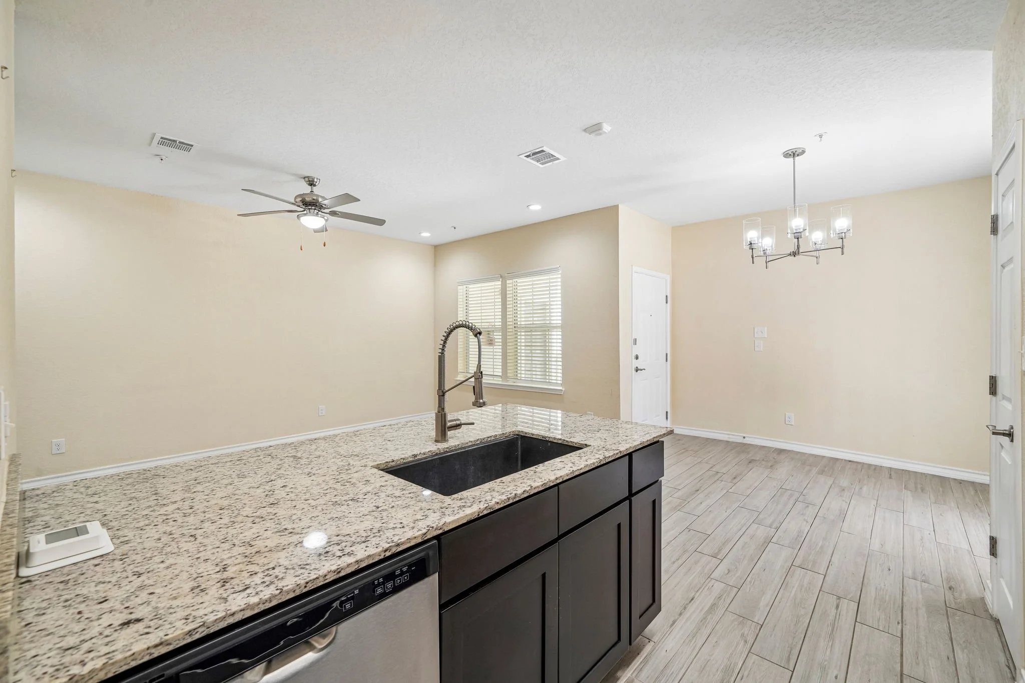 Open-concept kitchen with granite countertop island, stainless steel sink and faucet, beige walls, light wood flooring, ceiling fan, and a chandelier in the dining area, with windows and door in the background.