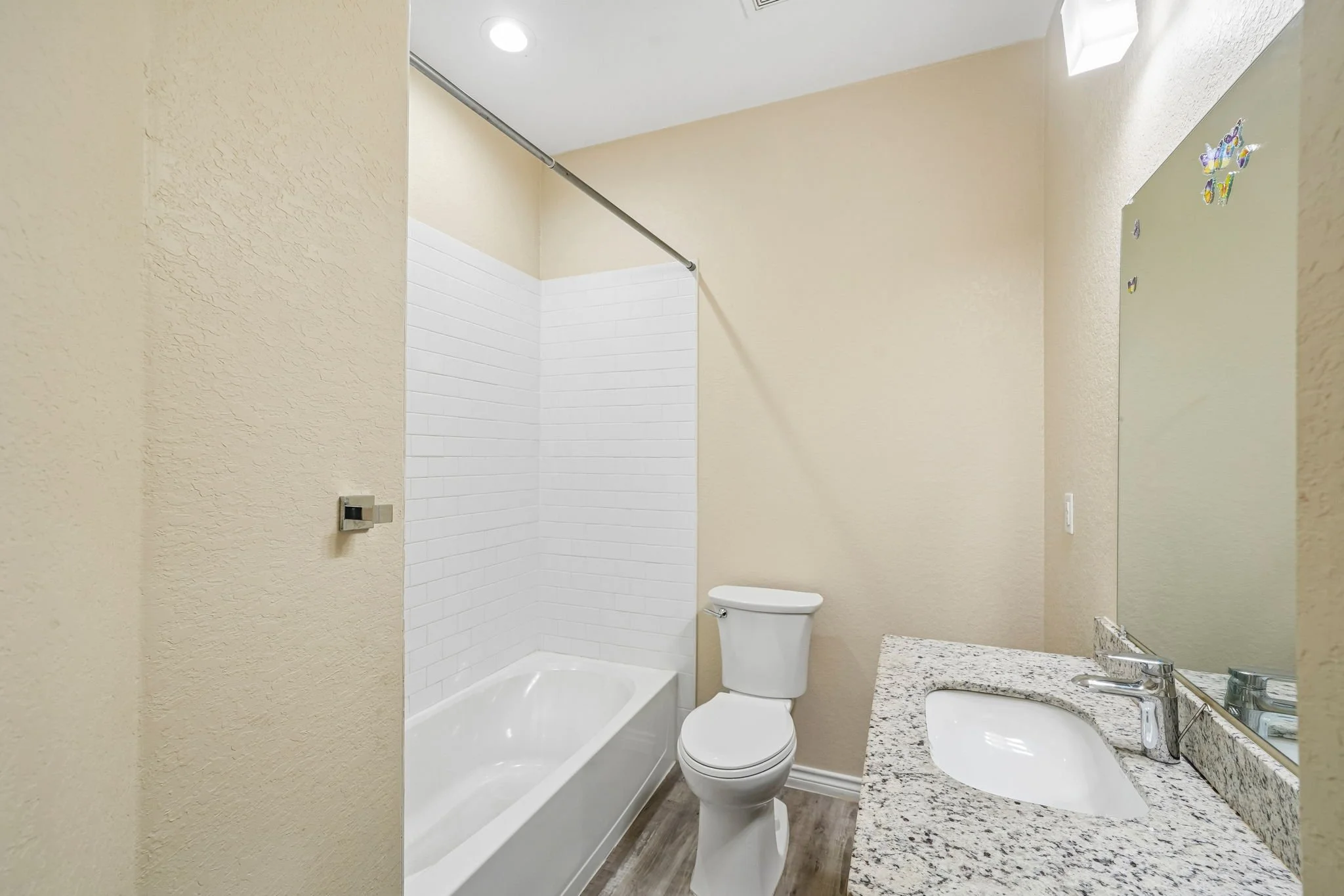 A small bathroom with beige textured walls, a white bathtub with a shower rod, a white toilet, a granite countertop with a sink, and a mirror with decorative stickers.