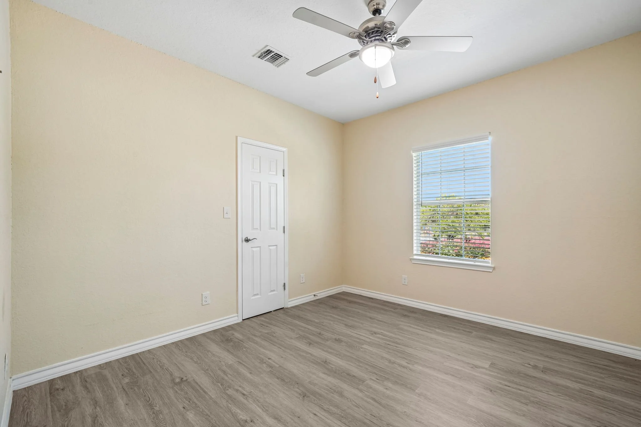 An empty bedroom with beige walls, a window with blinds, a ceiling fan, and laminate wood flooring.