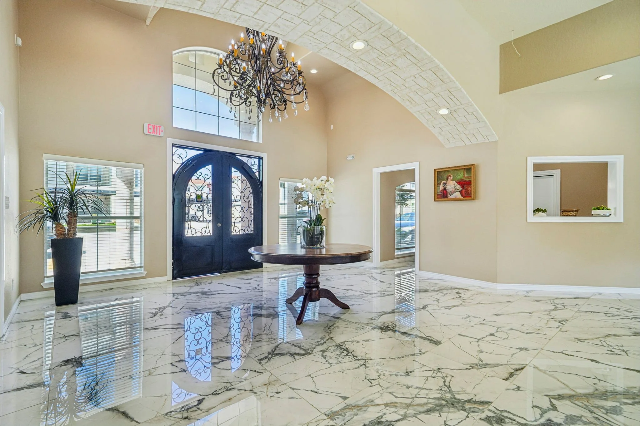 The elegant foyer of the La Marquesa Apartments clubhouse with a marble floor, a black front door with decorative ironwork, large windows, and a classic chandelier hanging from a vaulted ceiling.
